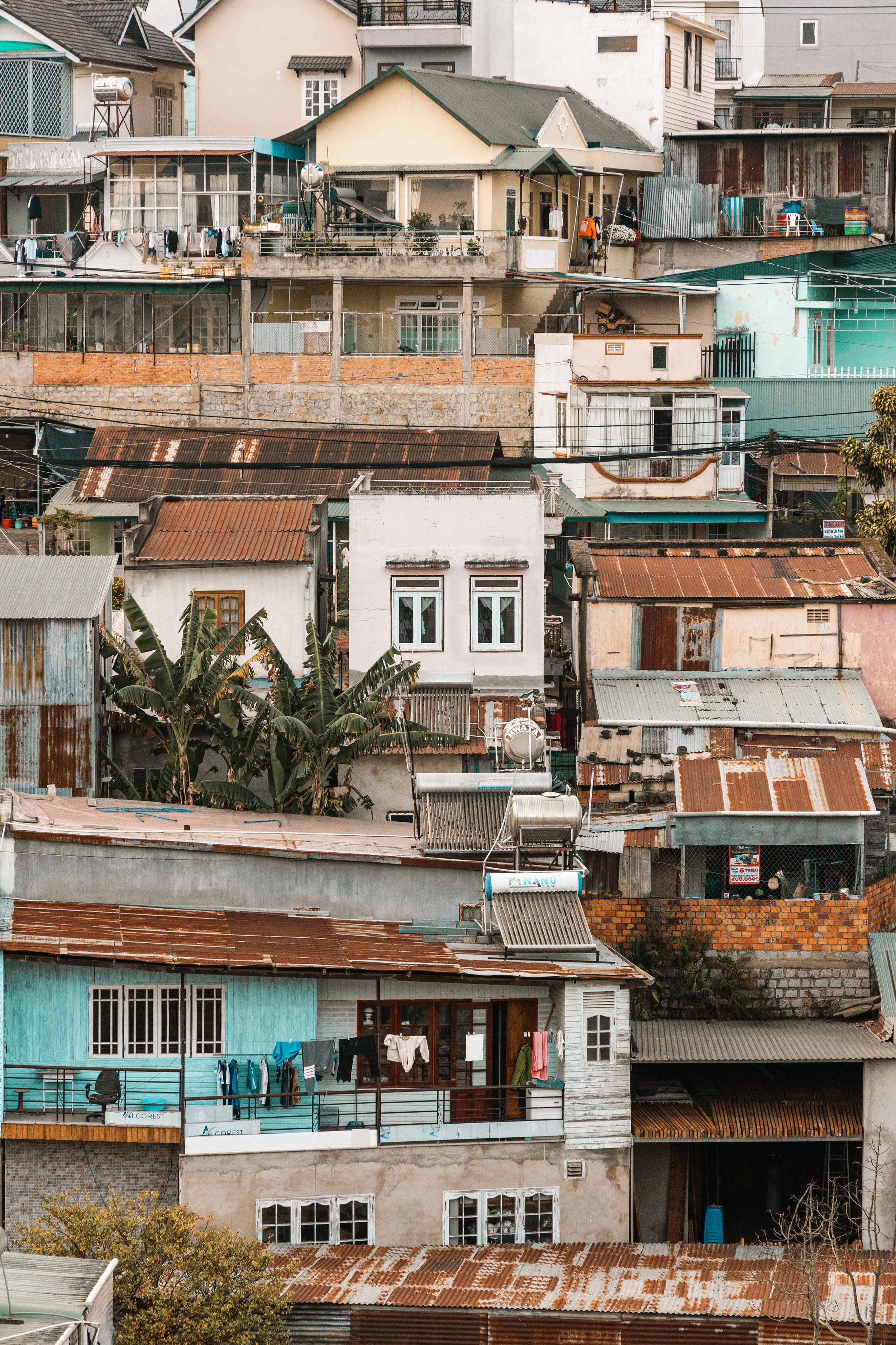 A densely packed hillside neighborhood with various colorful houses and buildings, some with metal and tile roofs, laundry hanging on balconies, and plants growing among the structures.