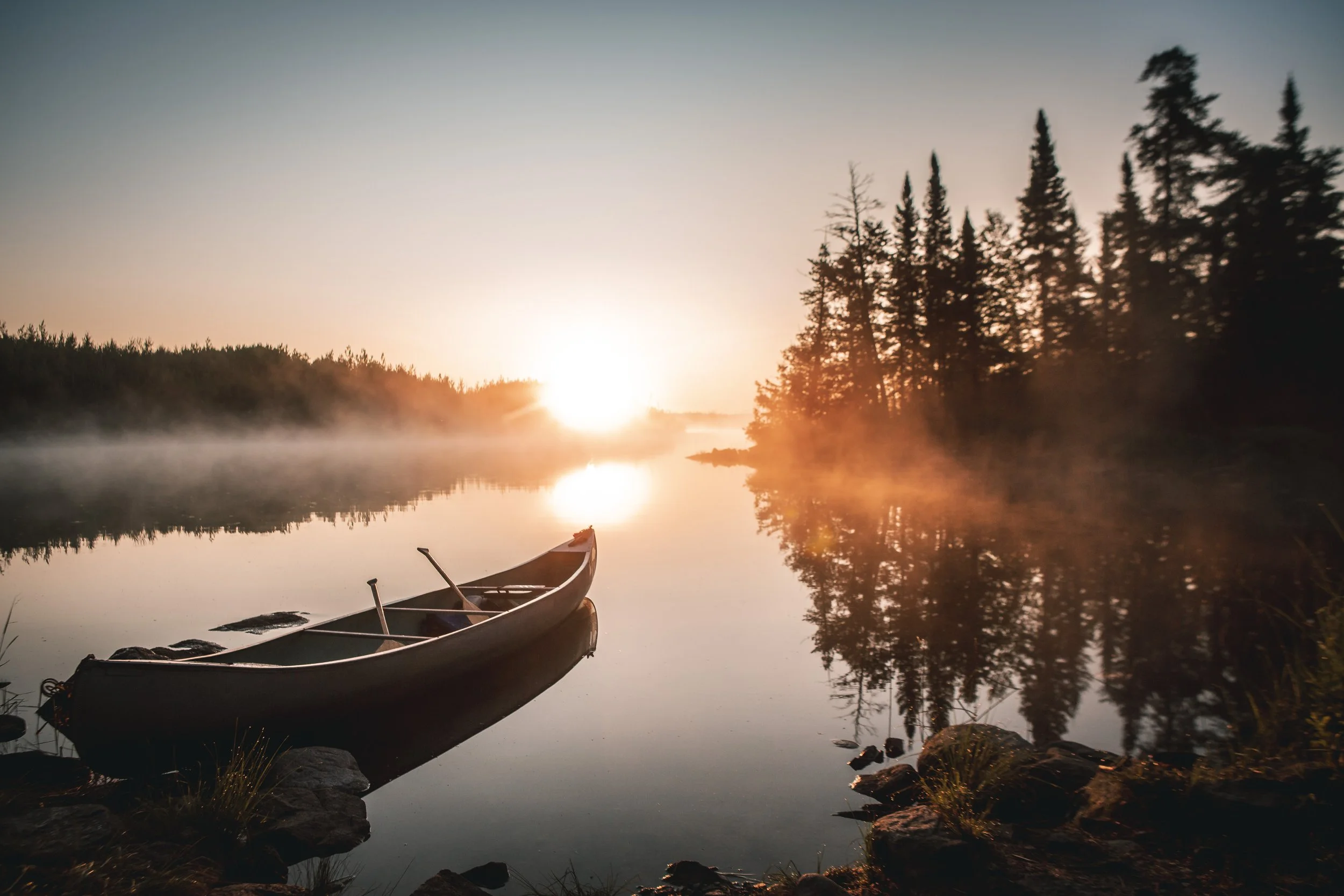 A peaceful lake scene with a canoe docked near the rocky shore during sunrise, surrounded by trees with mist rising from the water.