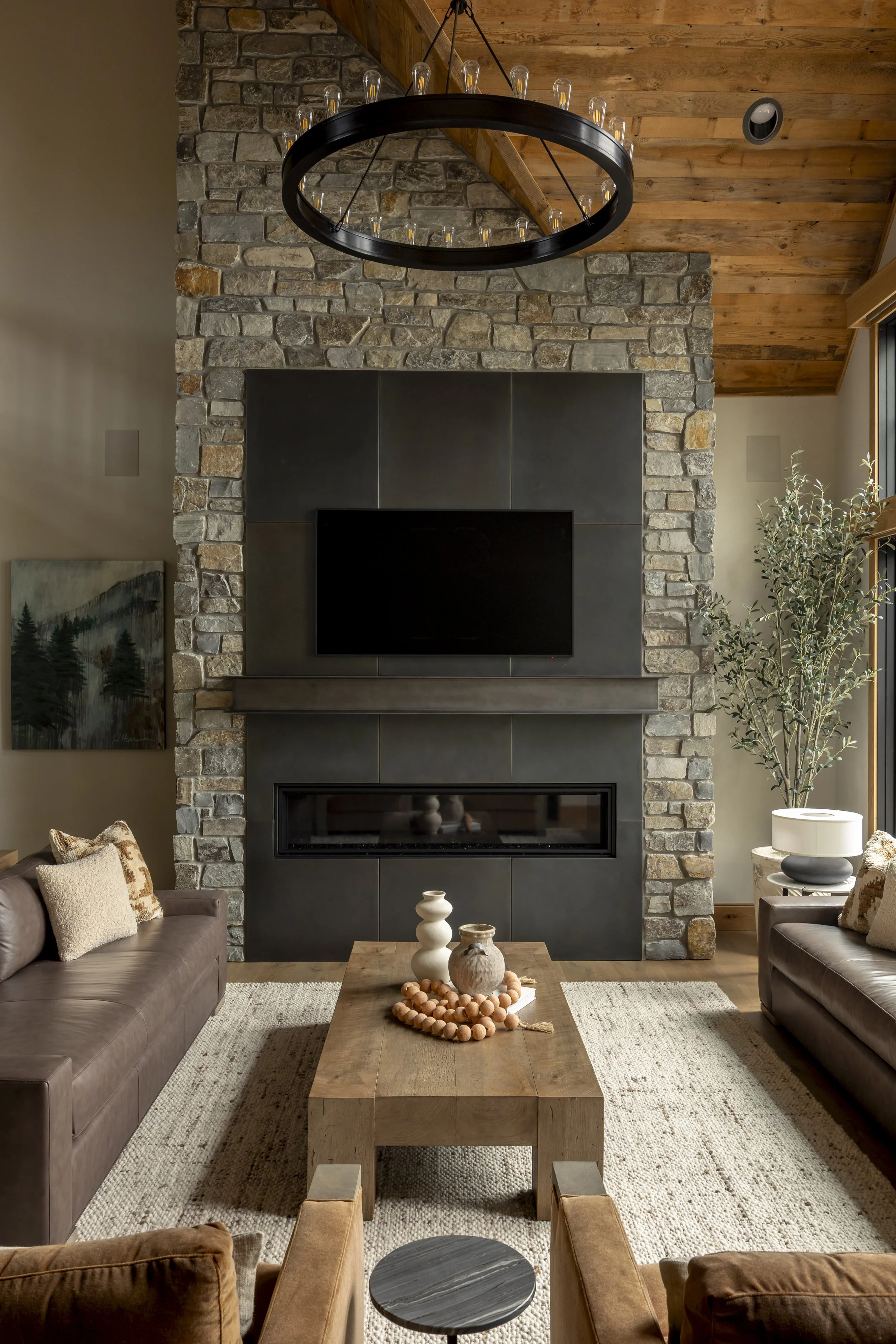 Living room with a tall brick fireplace, mounted flat-screen TV, wooden coffee table with decorative vases, and leather sofas. There's a black circular chandelier above and a large window with a potted plant on the side.