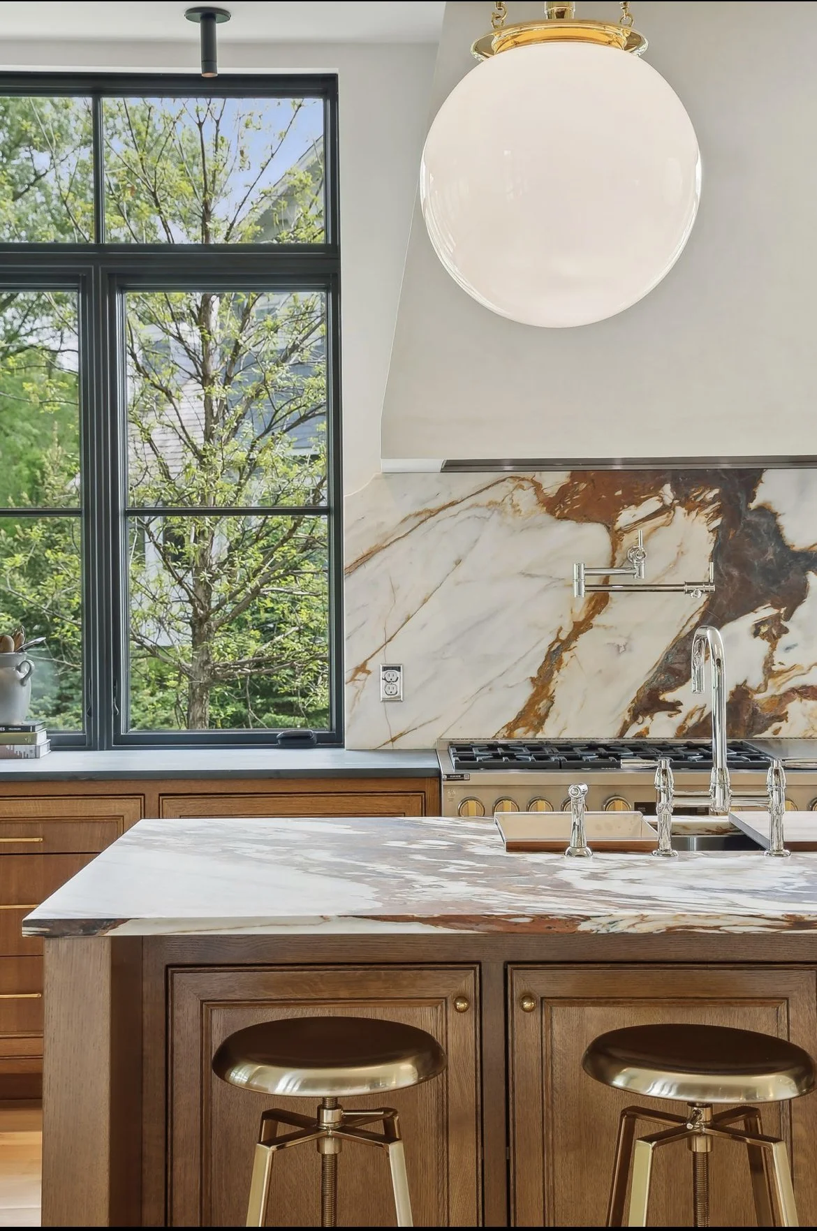 A modern kitchen with a large window showing green trees outside, a marble backsplash and countertop, wooden cabinetry, a large round white pendant light, and two metallic barstools at the island.