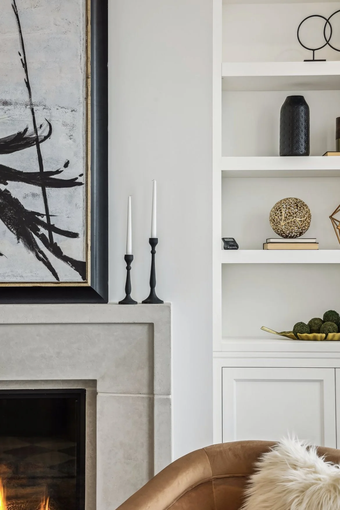 Close-up of a modern living room corner featuring a gray stone fireplace, two black and white candles on the mantel, and a white built-in shelf with decorative vases, books, and art.”}