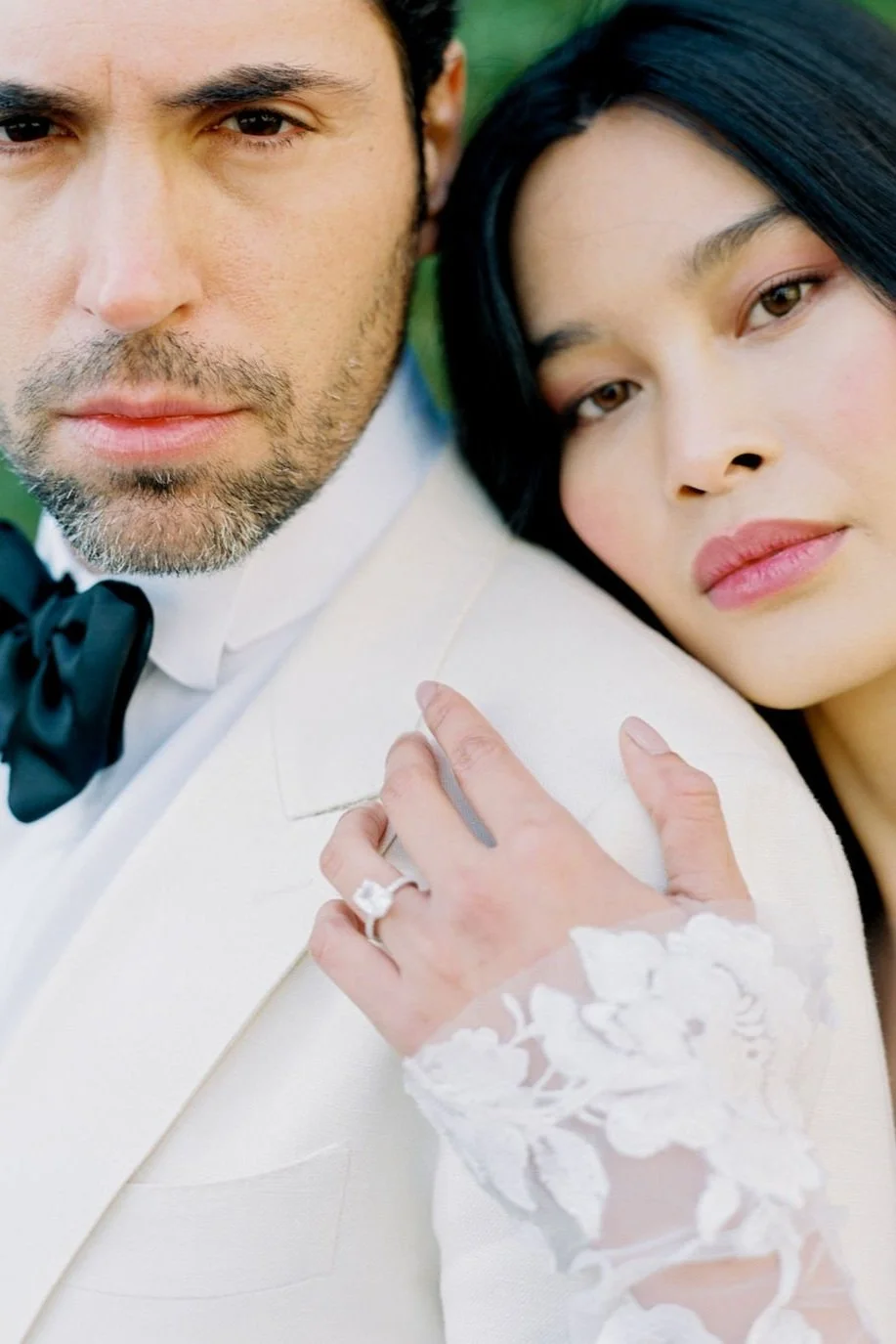 Close-up of a man with a beard and a woman with dark hair, both looking at the camera. The woman has her hand resting on the man's shoulder, showing a wedding ring. The man wears a white tuxedo with a black bow tie, and the woman wears a dress with lace sleeves.