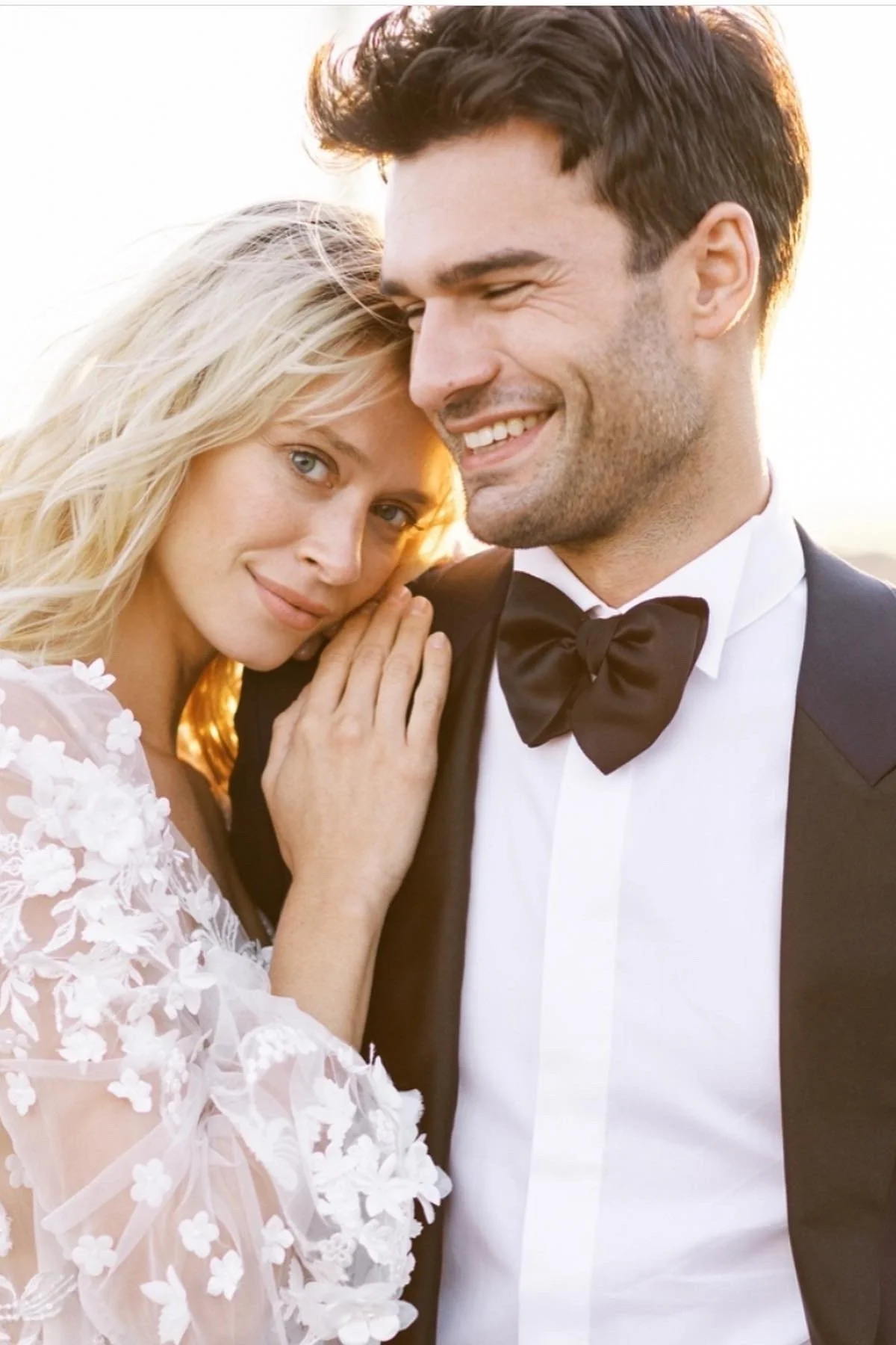 A smiling bride and groom on their wedding day during sunset, with the bride resting her head on the groom's shoulder.