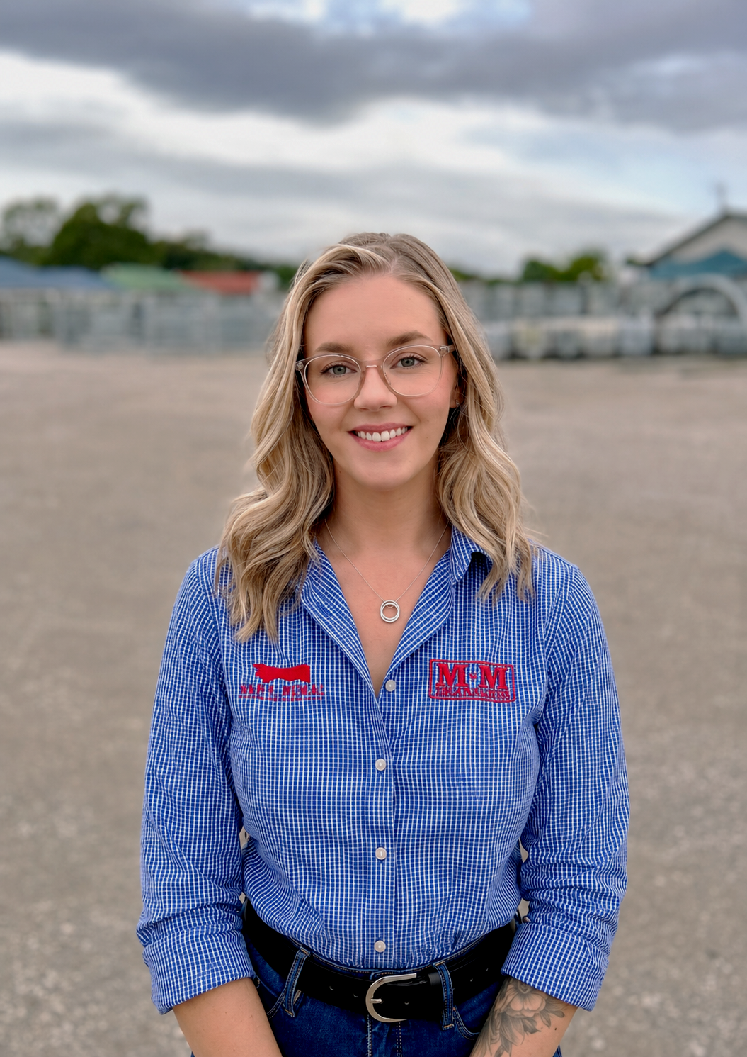 Young woman with glasses and tattoos wearing a blue checkered shirt with logos on it, standing outdoors in front of metal fencing.