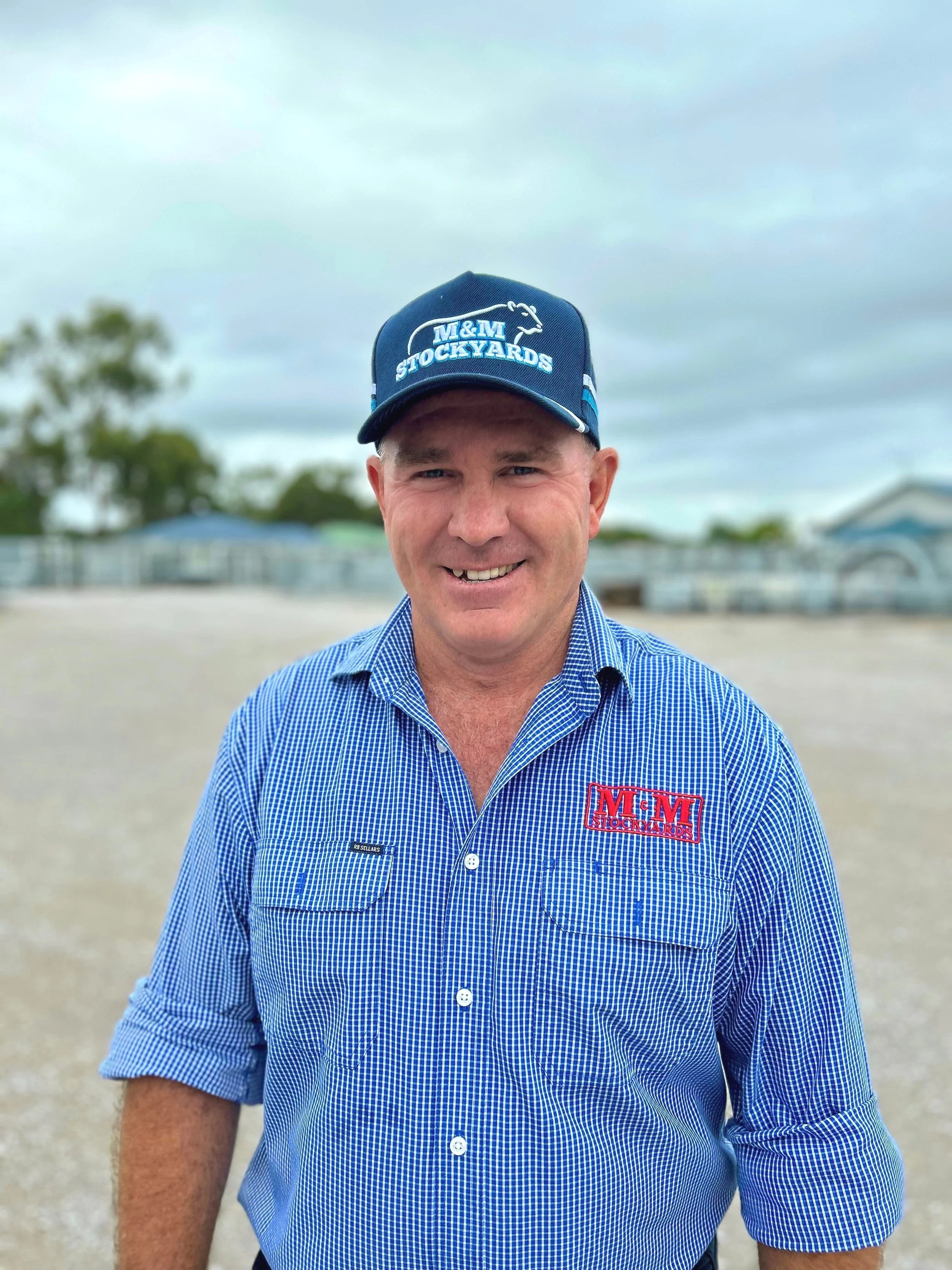 A man wearing a blue plaid button-up shirt with a logo and a blue cap with white and light blue text and a cattle image, standing outdoors on a gravel surface with trees and buildings in the background under a cloudy sky.
