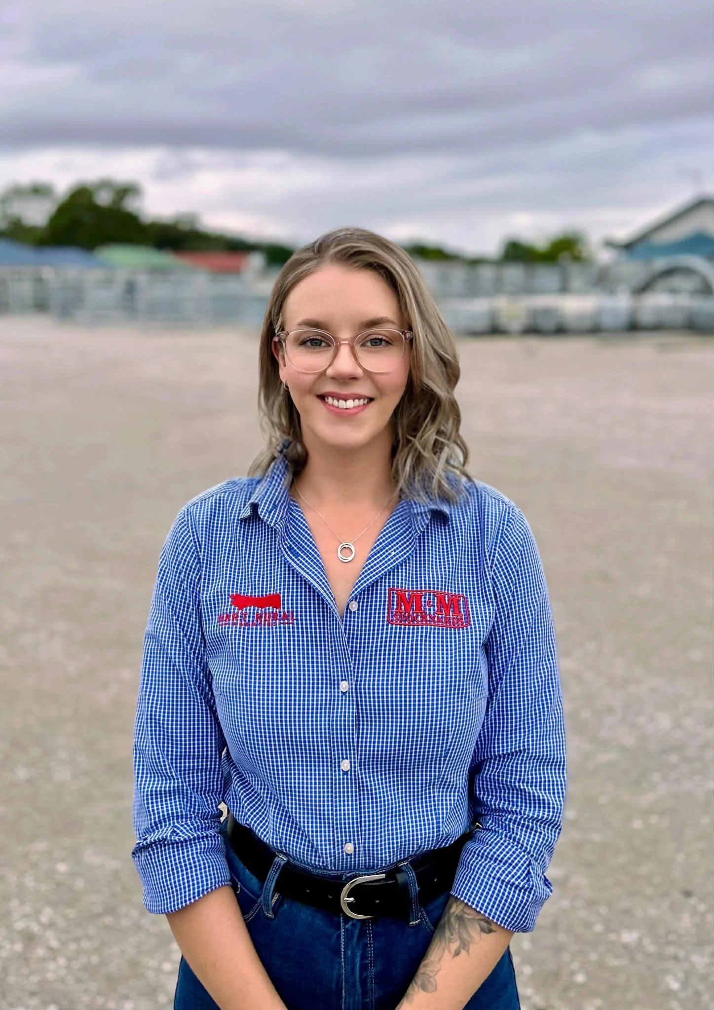 Young woman with glasses and tattoos wearing a blue checkered shirt with logos on it, standing outdoors in front of metal fencing.