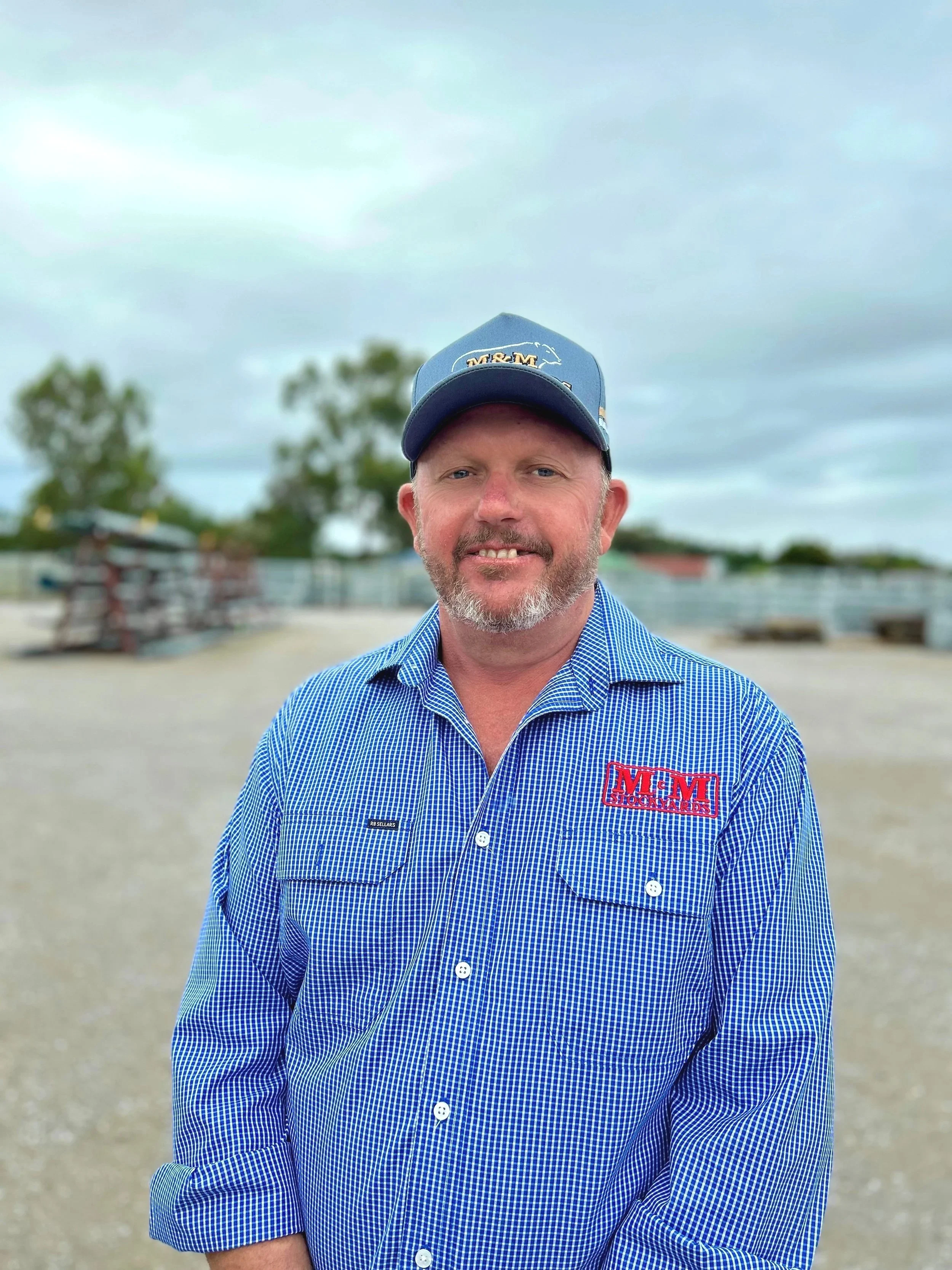 A man with a beard wearing a blue checkered shirt with red M&M graphics on the chest, and a blue cap, standing outdoors against a cloudy sky with trees and structures in the background.