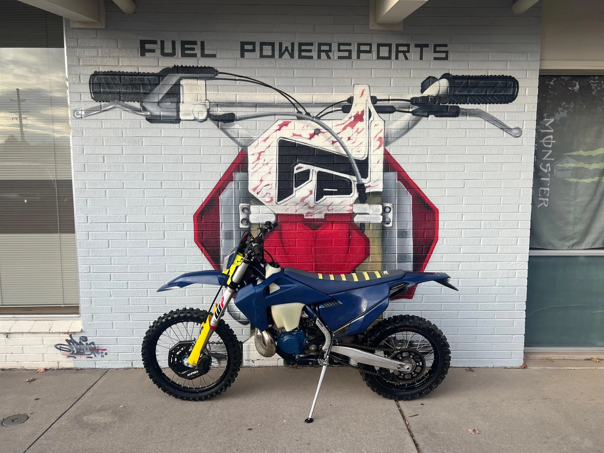 Off-road motorcycle parked in front of a large mural of a motorcycle handlebar on a brick wall, with the text 'FUEL POWERSports' above.