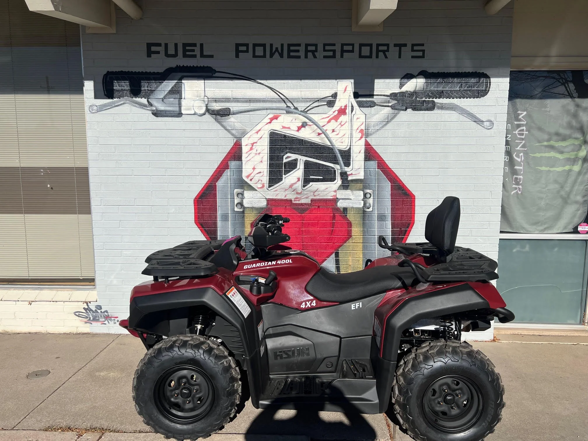 Red and black four-wheel all-terrain vehicle (ATV) parked in front of a painted wall that features a graphic of a motorcycle handlebar, grips, and a logo, with 'FUEL POWERSPORTS' painted above.