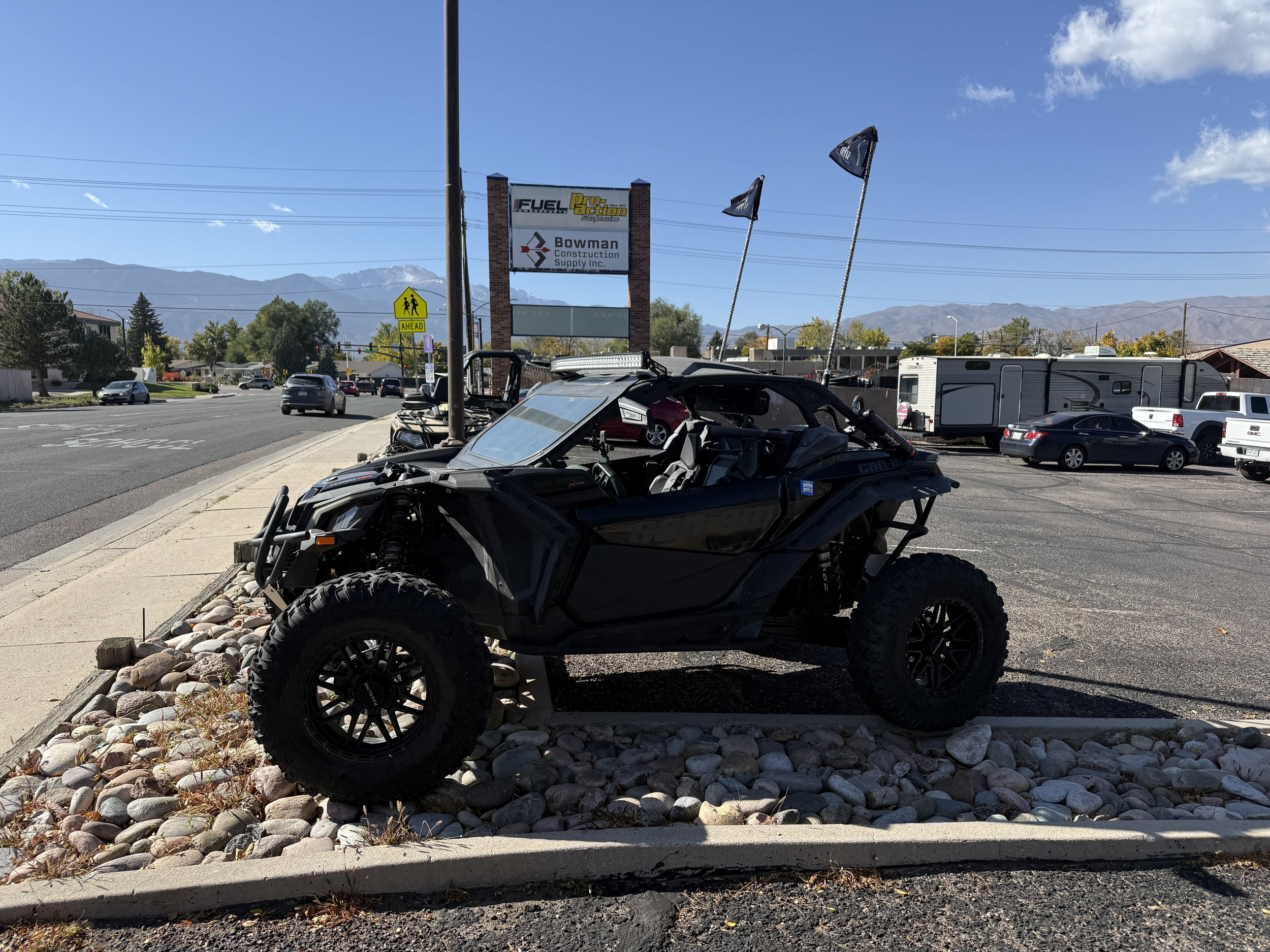 Black off-road vehicle parked on a gravel area beside a sidewalk in a parking lot with mountains in the background and a blue sky.