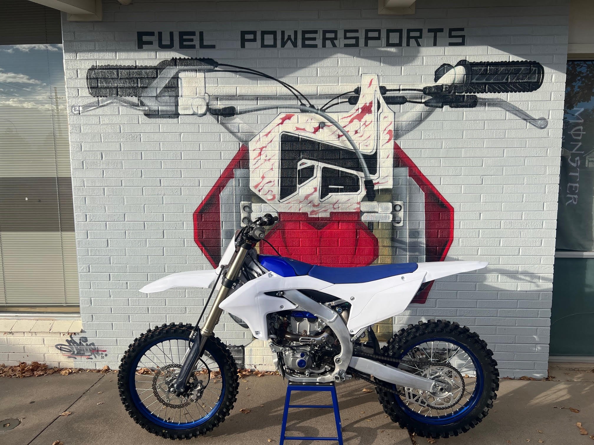 White and blue dirt bike motorcycle parked in front of a painted brick wall with a red and black motorcross helmet graphic and the words "Fuel Powersports" at the top.