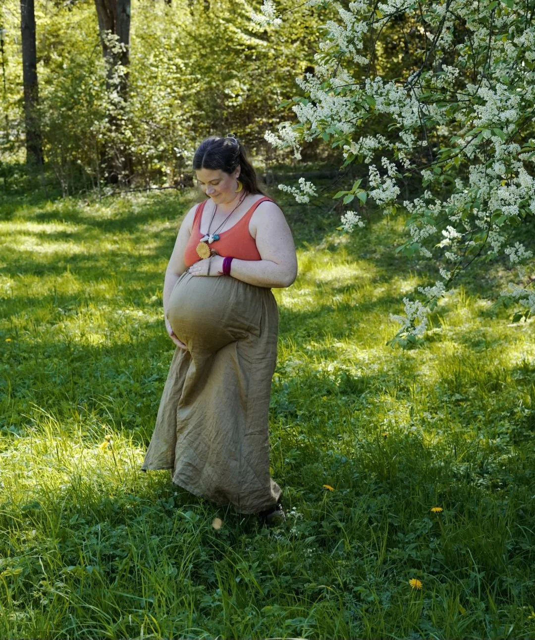 Gravid kvinna i en grönskande skogsmiljö med blommande buskar, bär på sin mage och ler.