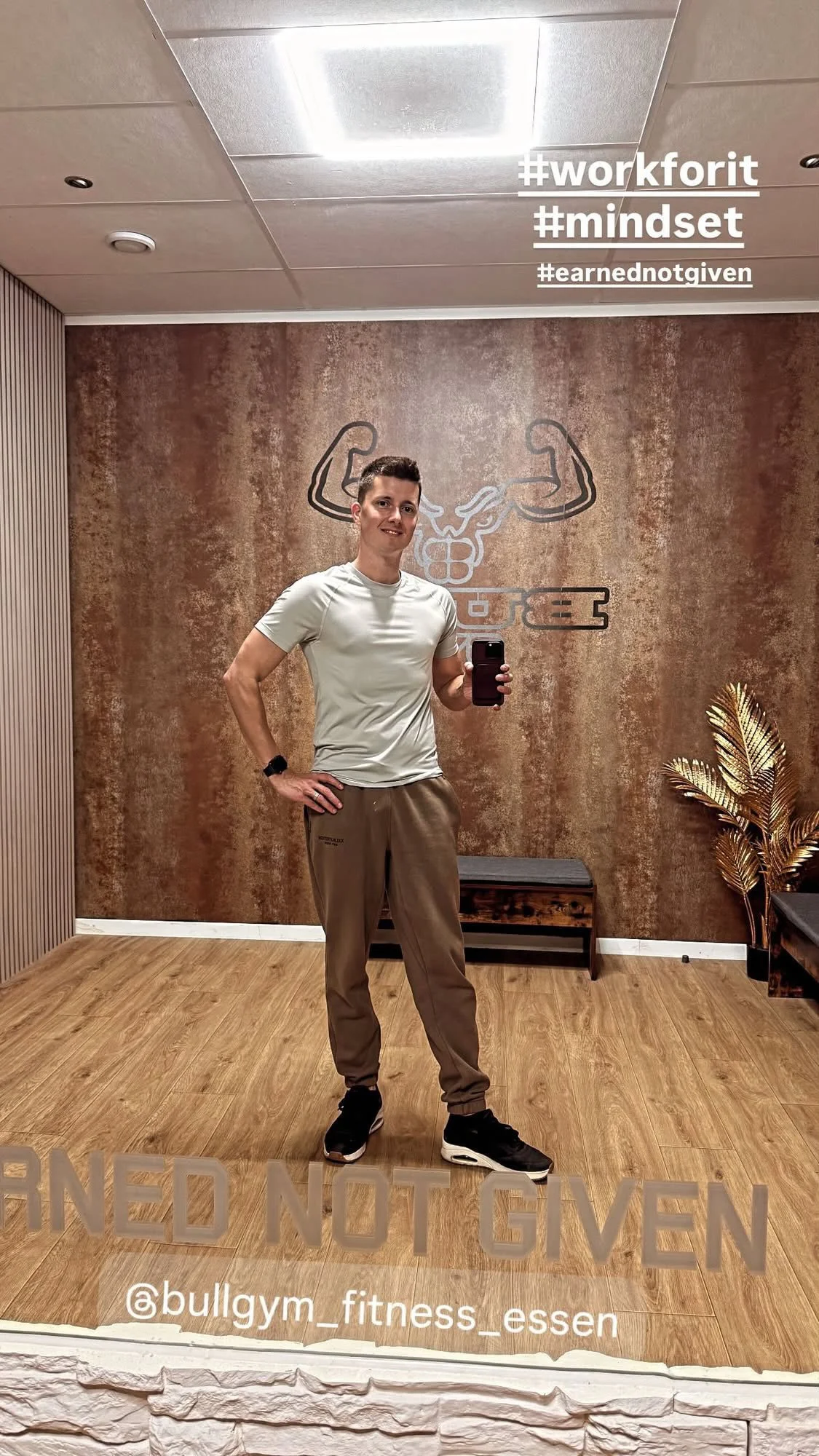 Man staat in een fitnessruimte, poseert voor een spiegel, draagt sportkleding, en er staan hashtags zoals #workforit, #mindset en #earnednotgiven in de hoek.