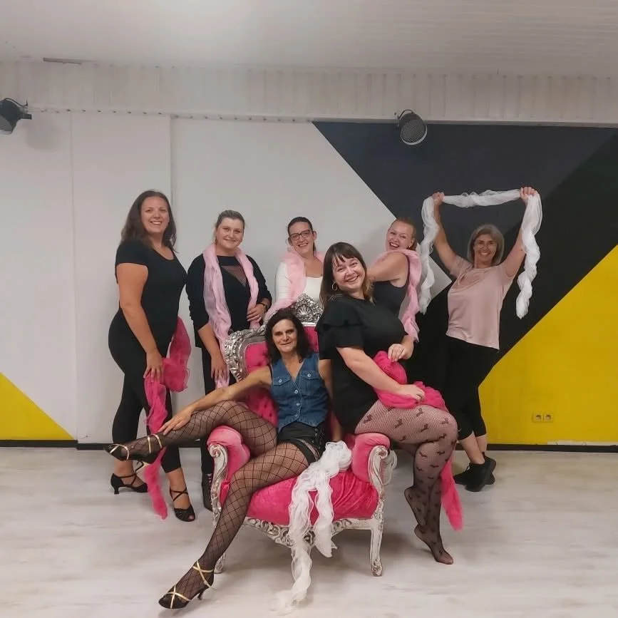 Zeven vrouwen van burleske dansen.  poseren in een kamer, met één vrouw zittend op een roze en zilveren troon en de andere zes vrouwen eromheen. Ze lachen en houden linten en sjaals vast, met een kleurrijke muur op de achtergrond.