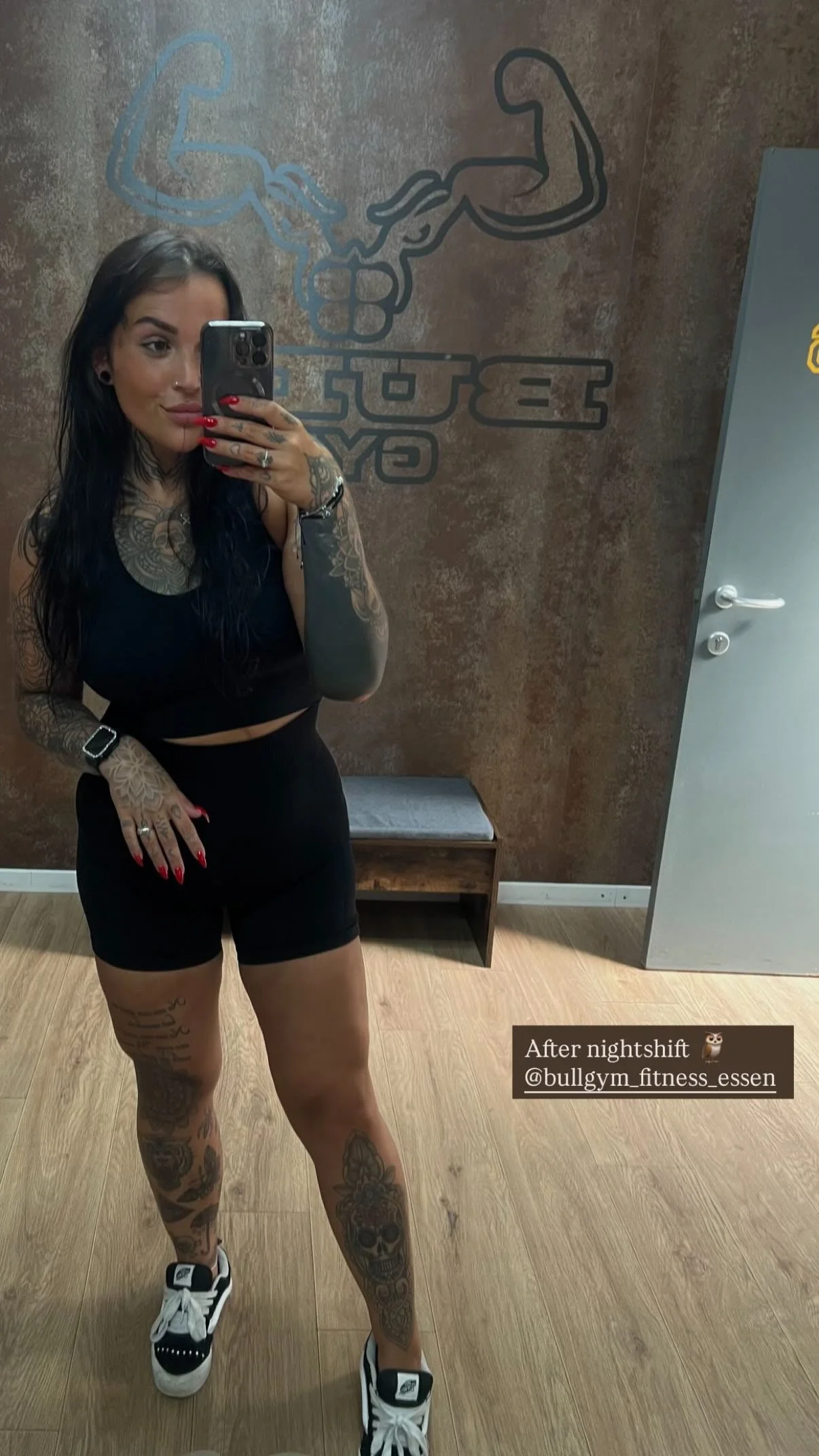 Een vrouw met tatoeages op armen en benen, gekleed in zwart sportoutfit en witte sneakers, neemt een selfie in de fitnessruimte. Op de muur achter haar een muurkunstwerk van biceps en een stang met gewichten. Tekst op de afbeelding: 'After nightshift @bullgym_fitness_essen'.