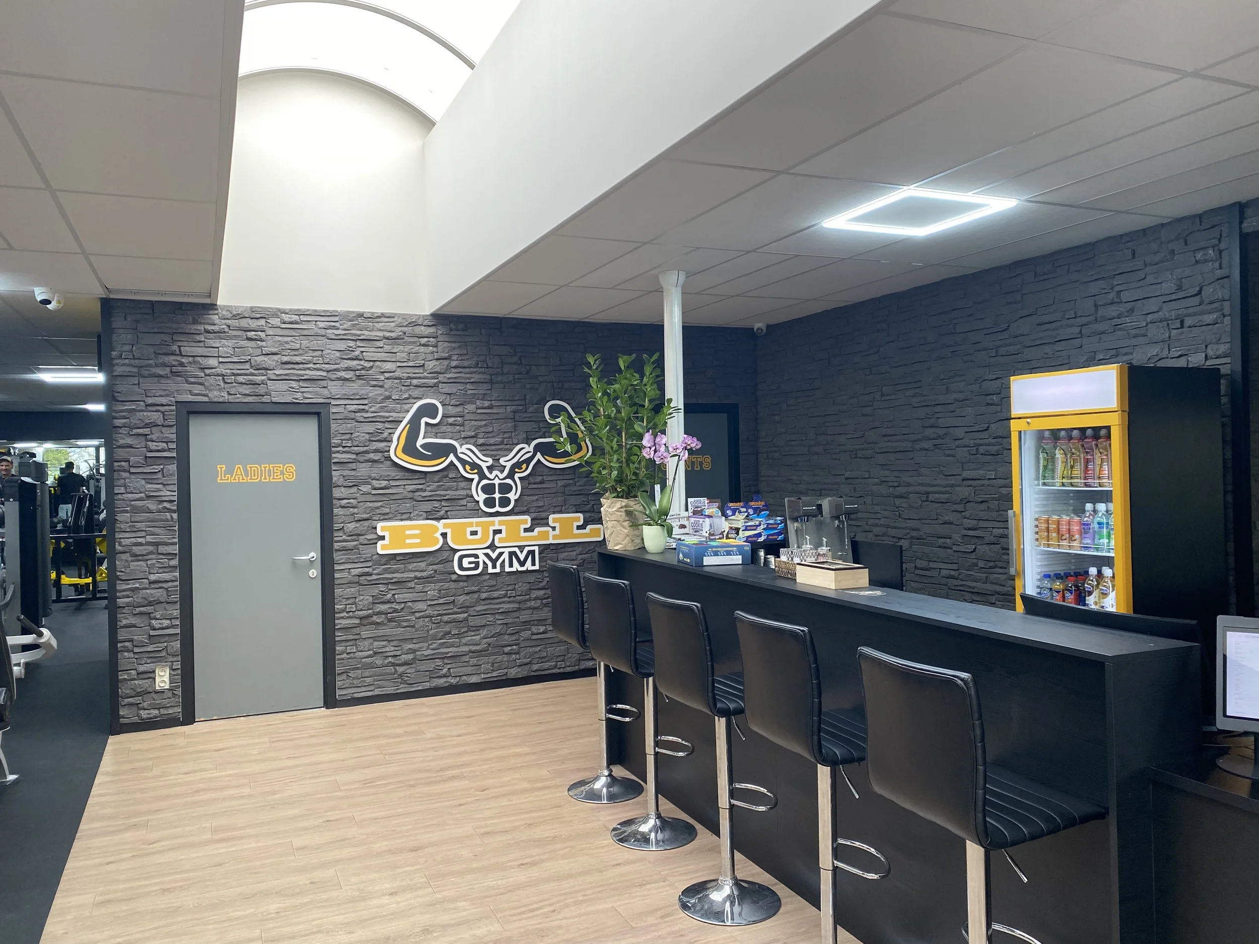 Binnenkant van een fitnesscentrum met een bar en zitplaatsen, fitnessapparatuur en gewichten op de achtergrond.