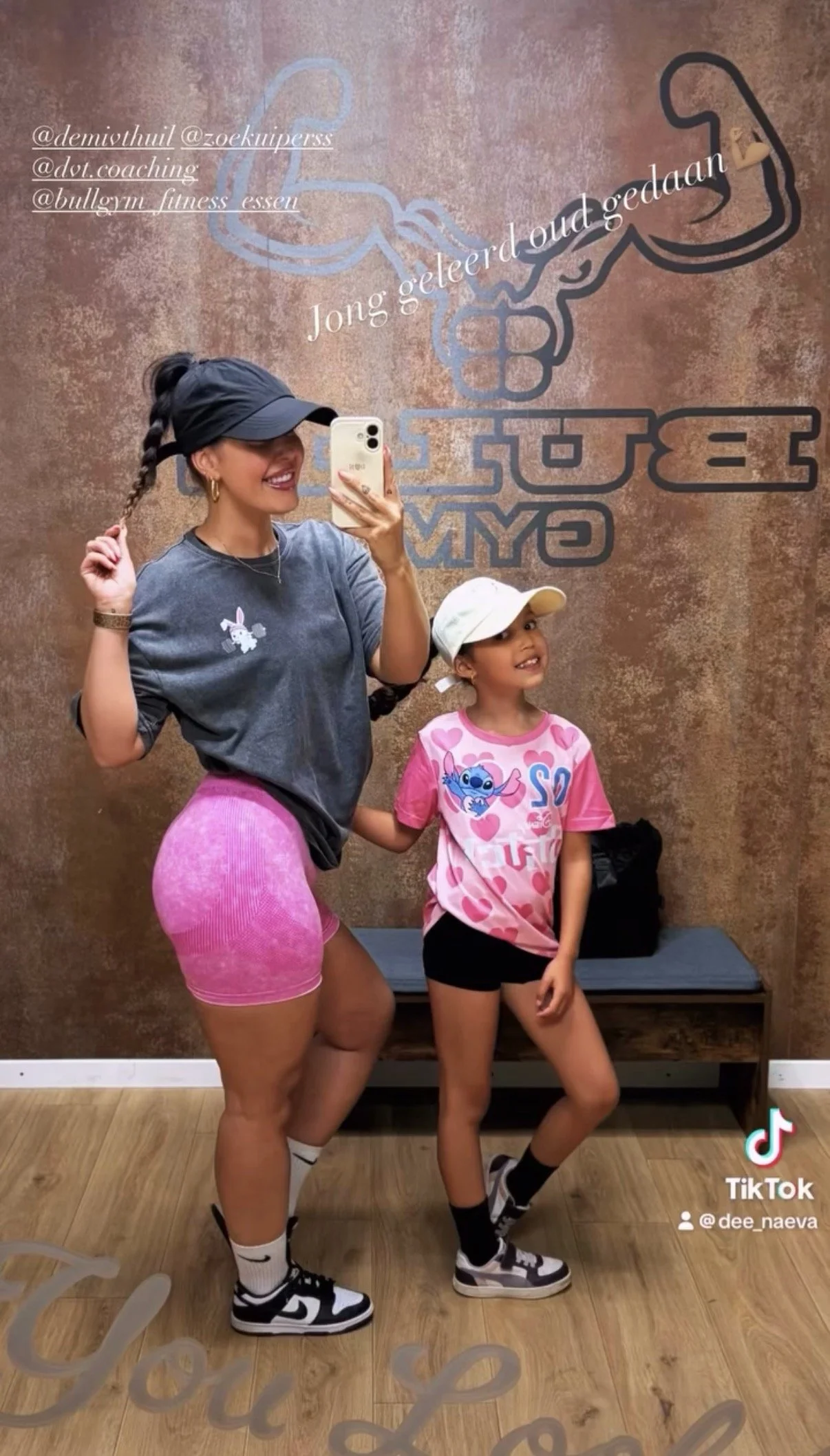 Twee meisjes poseren voor een spiegel, een glimlachend jonge vrouw met een piraatssjaal en roze sportbroek, en een klein meisje met een witte pet en een roze T-shirt met heart-toestanden. Ze nemen een selfie in het fitnesscentrum.