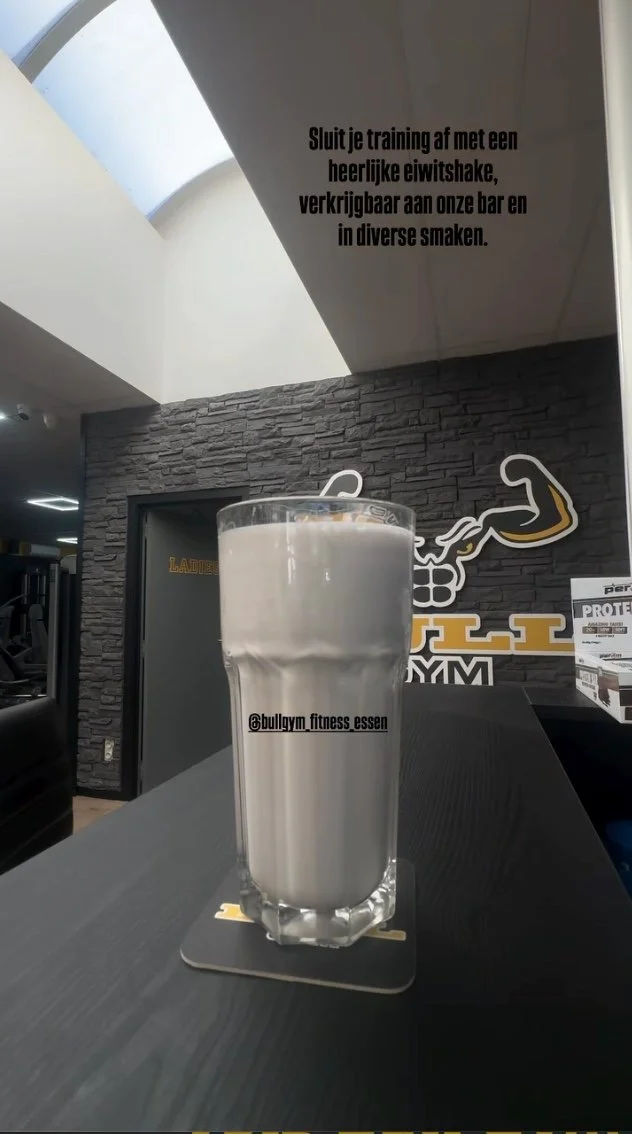 Glas met witte eiwitshake op een zwart oppervlak in een fitnessruimte met een logo op de muur op de achtergrond.