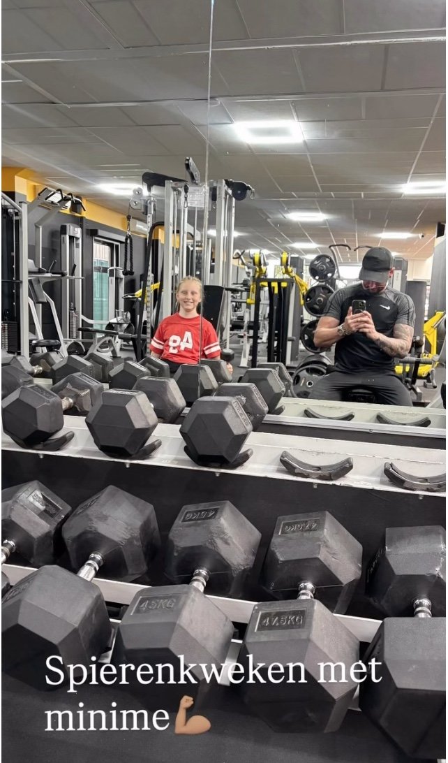 Binnenin een sportschool met een spiegel die rijen halters (gewichtheffers) reflecteert. Tegen de spiegel staan een meisje met een rode trui en een man met een zwart t-shirt en pet, die zich via de spiegel opnemen.