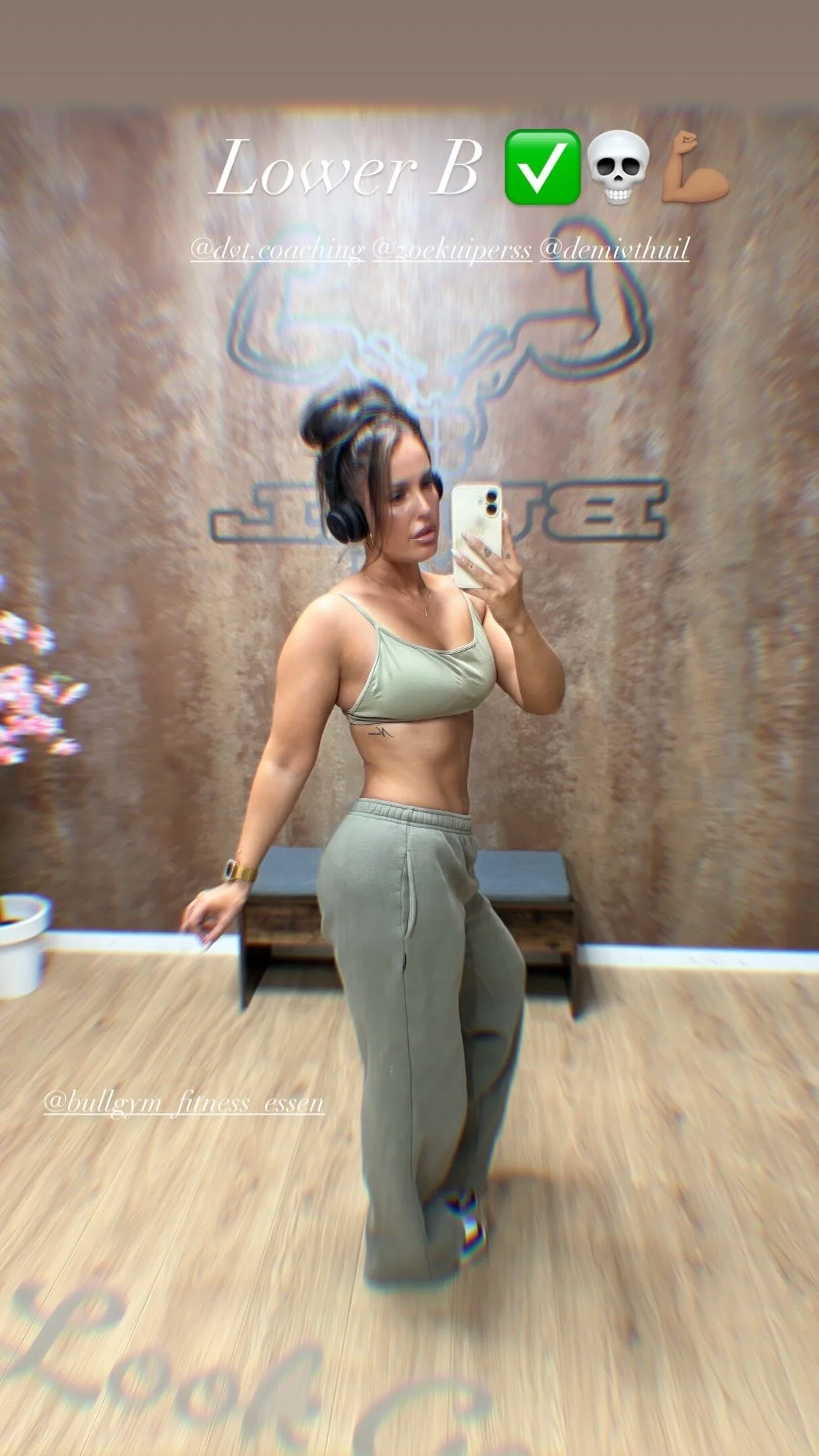 Een vrouw in sportkleding neemt een selfie in een spiegel, met een achtergrond van een muur met een logo en tekst.