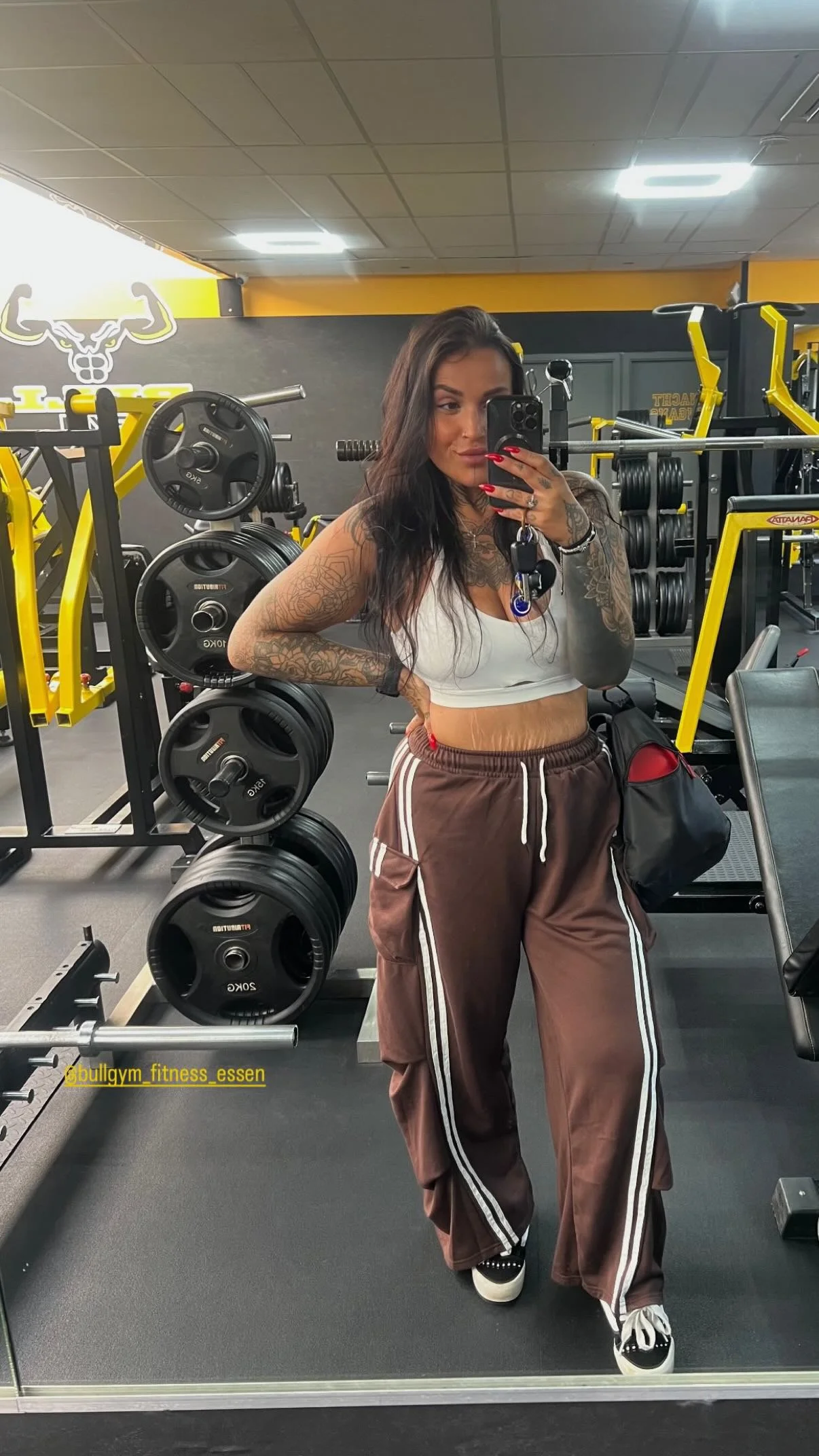 Een vrouwelijke persoon met tatoeages poseert voor een spiegel in een fitnessruimte, gekleed in een witte sporttop en bruine trainingsbroek met witte strepen, terwijl ze haar telefoon gebruikt voor een selfie. Fitnessapparatuur en gewichten zijn zichtbaar op de achtergrond.