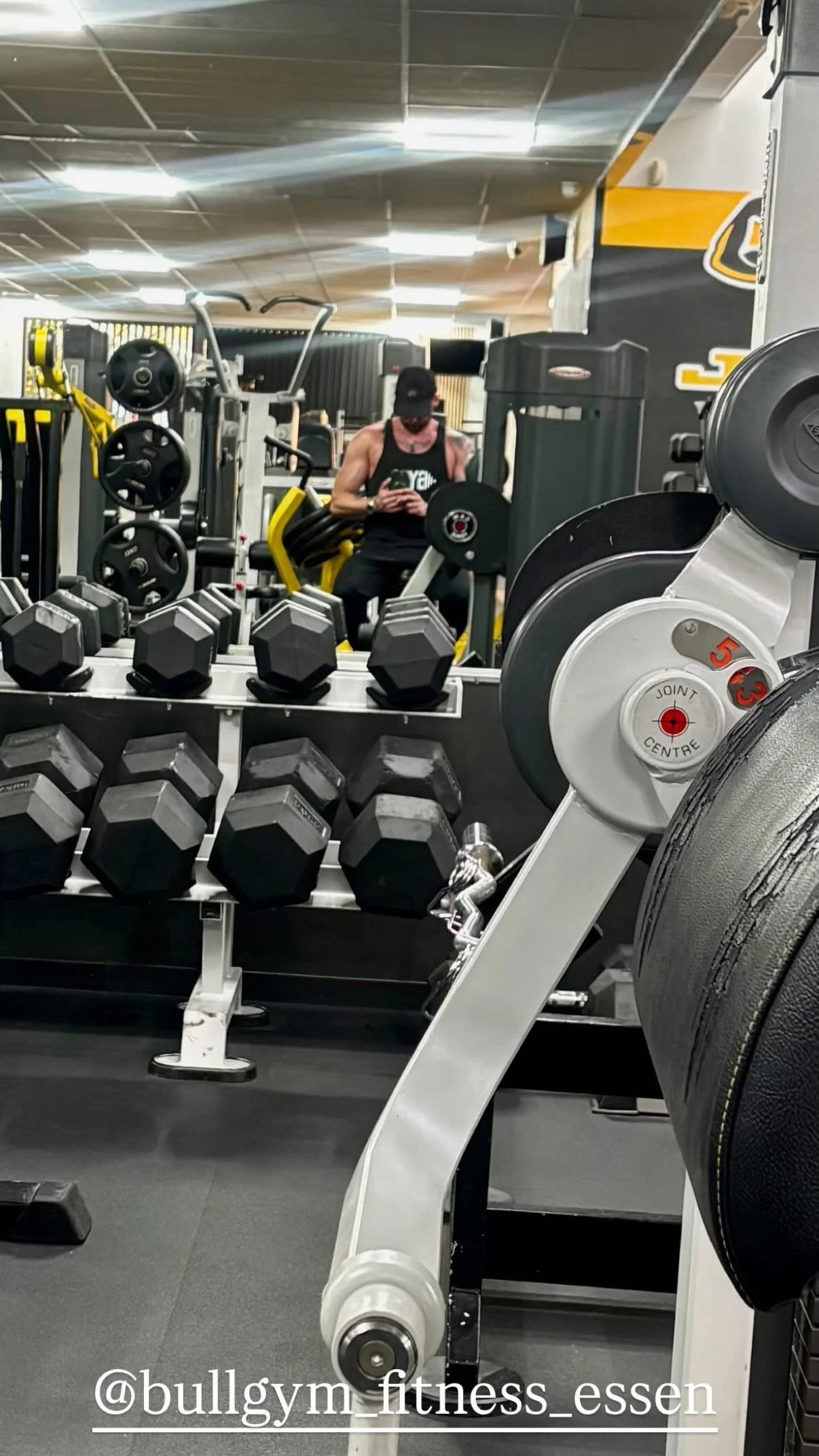 Een fitnessruimte met verschillende gewichten en fitnessapparaten, persoon met spiermassa in sportkleding, die in de spiegel kijkt.