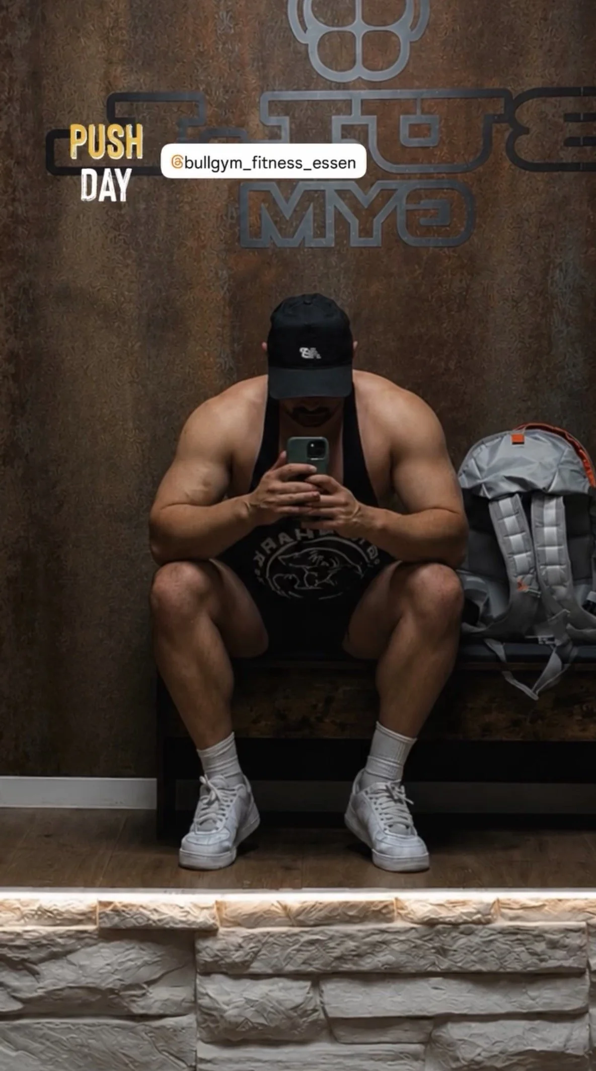 Man die in een fitnessruimte een spiegel besteedt, zittend op een bankje met een rugzak naast zich, terwijl hij een selfie neemt in de spiegel.