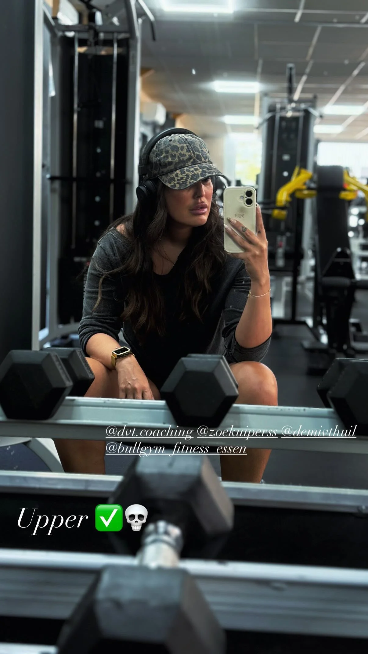 Een vrouw in sportkleding en een leopard print pet neemt een selfie in de spiegel van een sportschool, met gewichten en fitnessapparatuur op de voorgrond, en fitnessruimtes op de achtergrond.