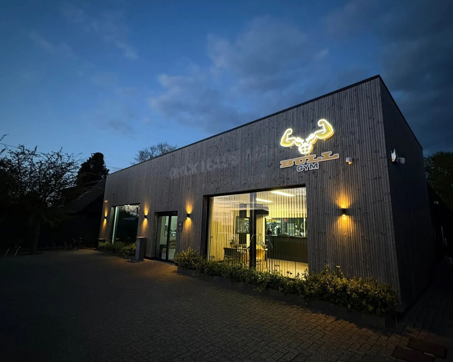 Een fitnesscentrum genaamd 'BULL GYM' 's avonds met een verlichte boksbal en spierarmlieten in het logo op de gevel, omgeven door buitenverlichting langs de muur.