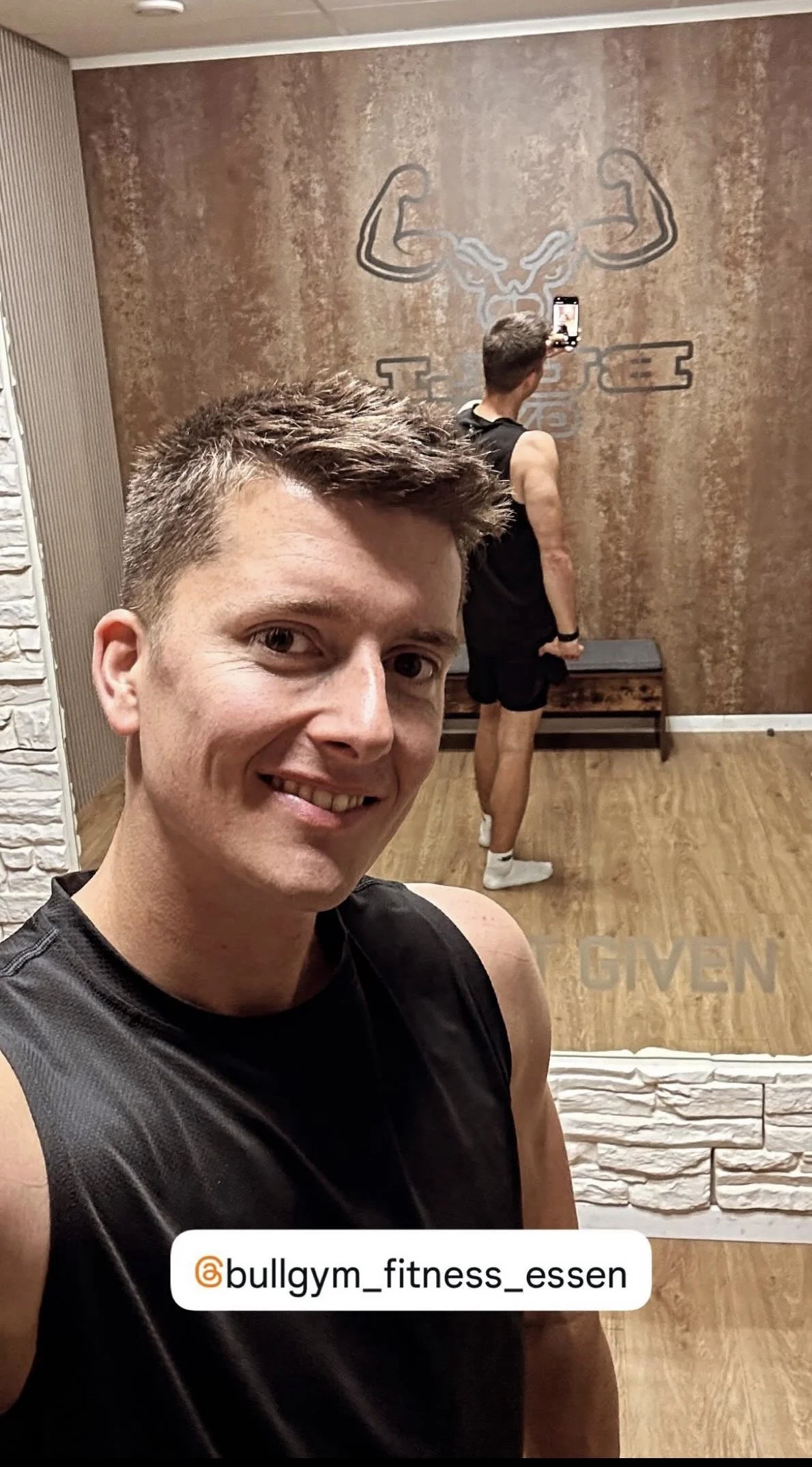 Een lachende man in een sportschool neemt een selfie. Achter hem staat een man met een sportbroek en een fitnesslogo op de muur.