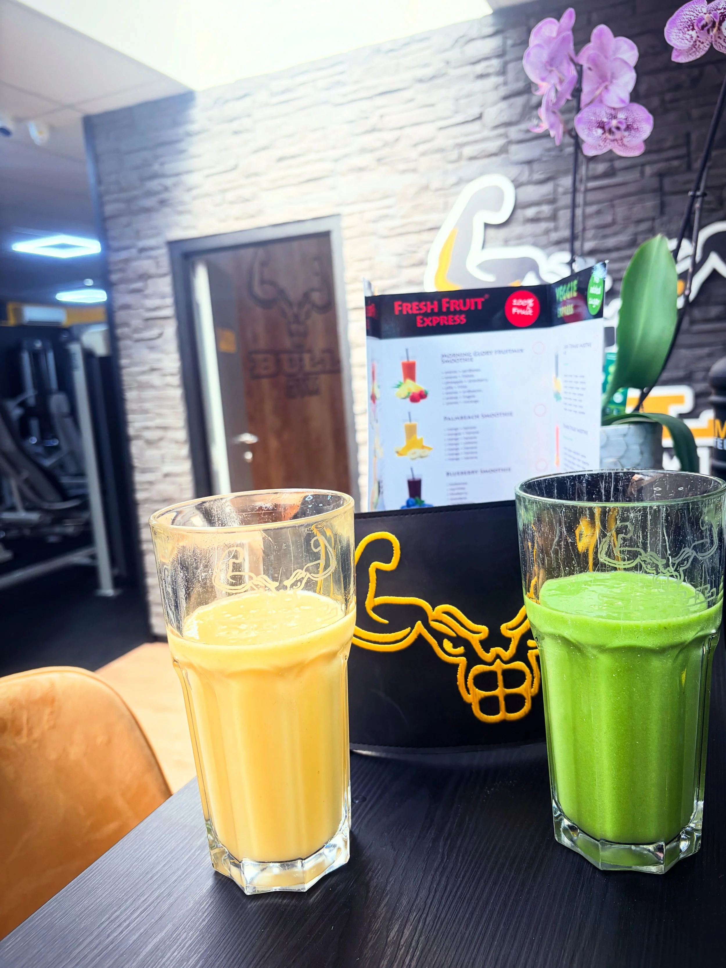 Twee glazen met smoothies, een gele en een groene, op een zwarte tafel. Op de achtergrond een menu en decoratieve bloemen, met een donker bakstenen muur.