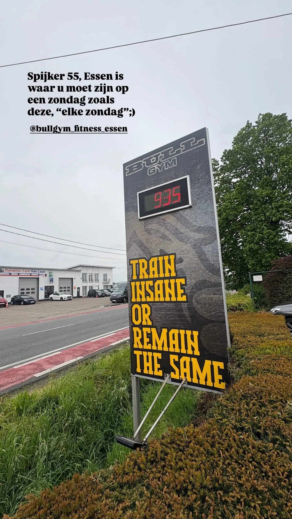 Sign with digital clock showing 9:35, text in Dutch over the top and yellow text on black background below, with a parking lot and trees in the background.