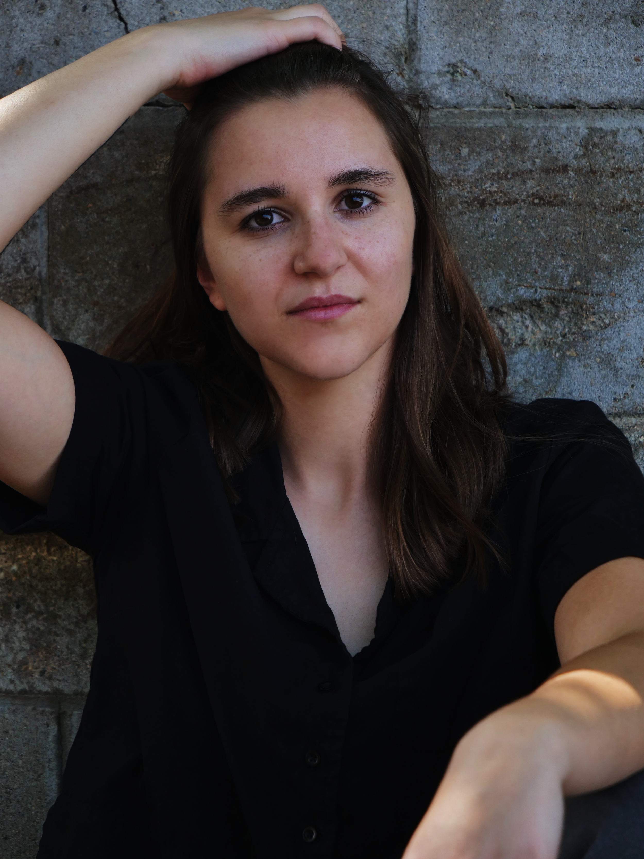 A young woman with shoulder length dark brown hair, dark brown eyes, wearing a black button-up short-sleeve shirt, is pushing her hair back with her right hand, standing against a stone wall background.