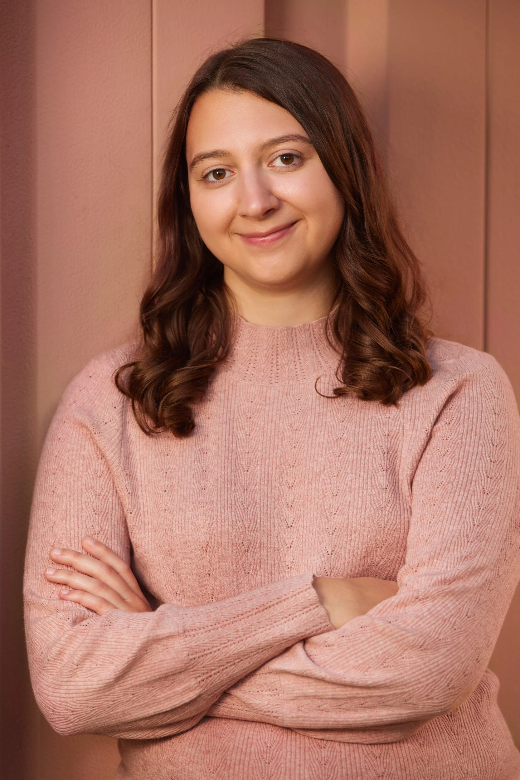 A smiling young woman with shoulder-length, curly brown hair, and brown eyes, wearing a pink knitted sweater stands in front of a neutral background.