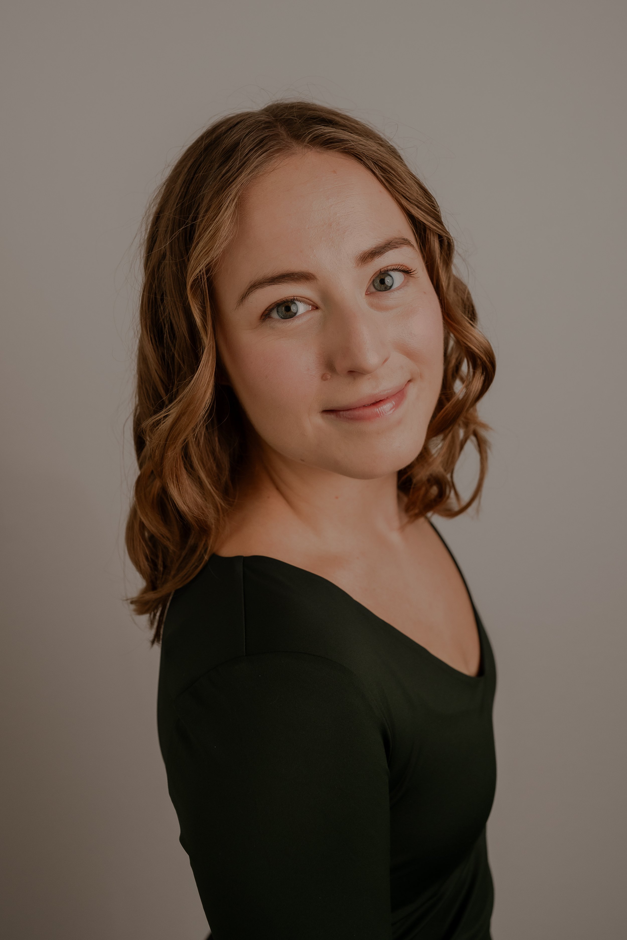 A young woman with green eyes and light brown hair, wearing a black long sleeve v-neck is smiling and standing in front of a neutral background
