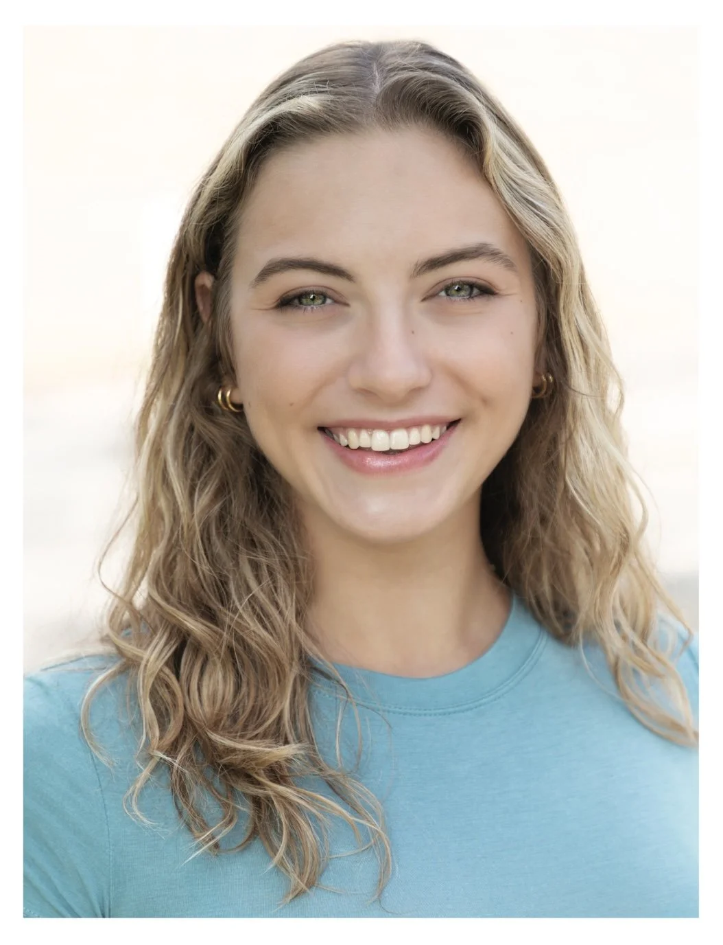A young woman with curly blonde hair and blue eyes wearing a blue shirt is smiling in front of a white background.