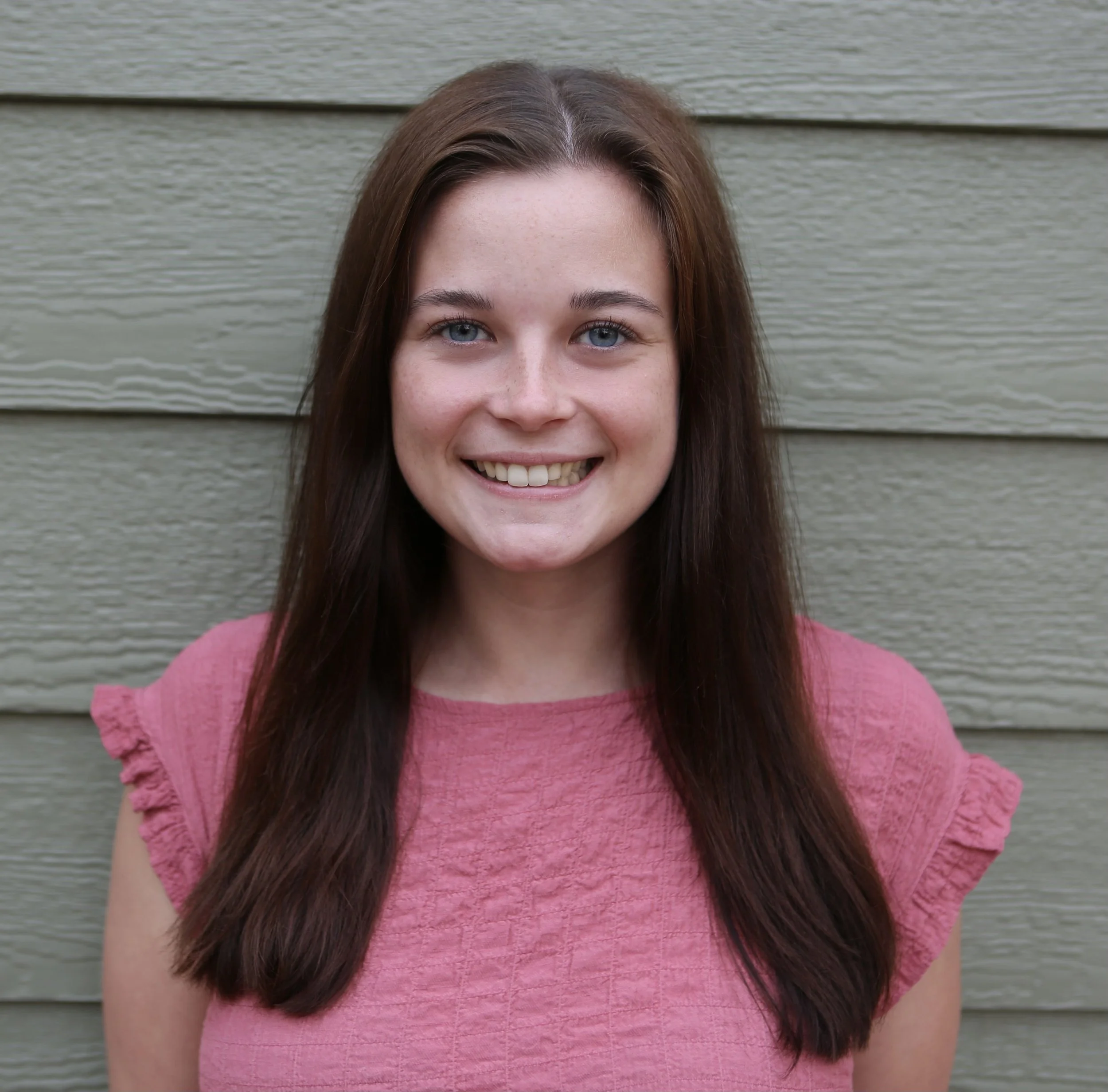 A young woman with long dark hair, blue eyes, and a big smile, wearing a textured pink shirt with pleated sleeves is standing in front of a neutral background.