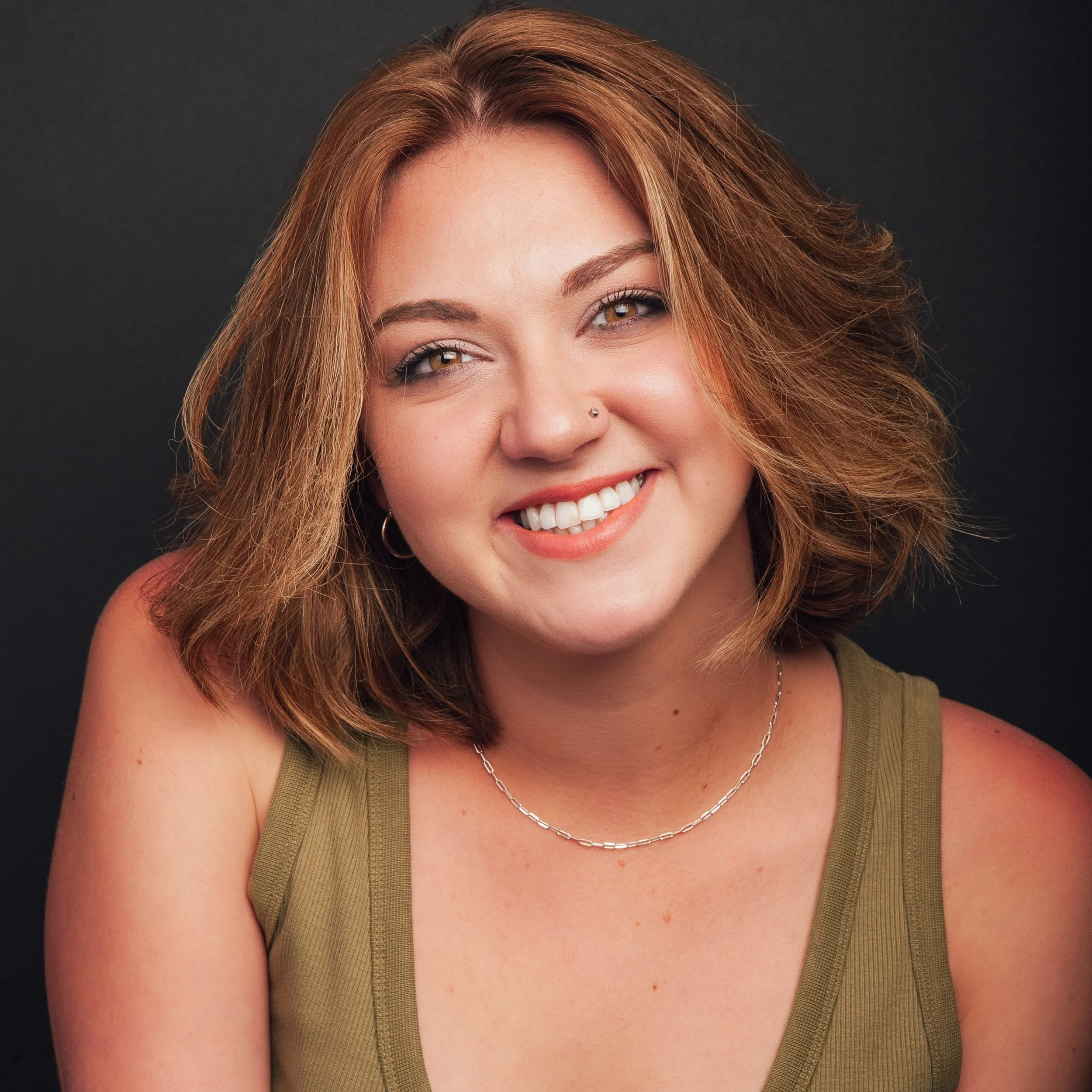 A young woman with shoulder-length light brown hair, amber eyes, and a bright smile, wearing a green tank top, a silver chain necklace, and a nose stud, standing against a neutral background.