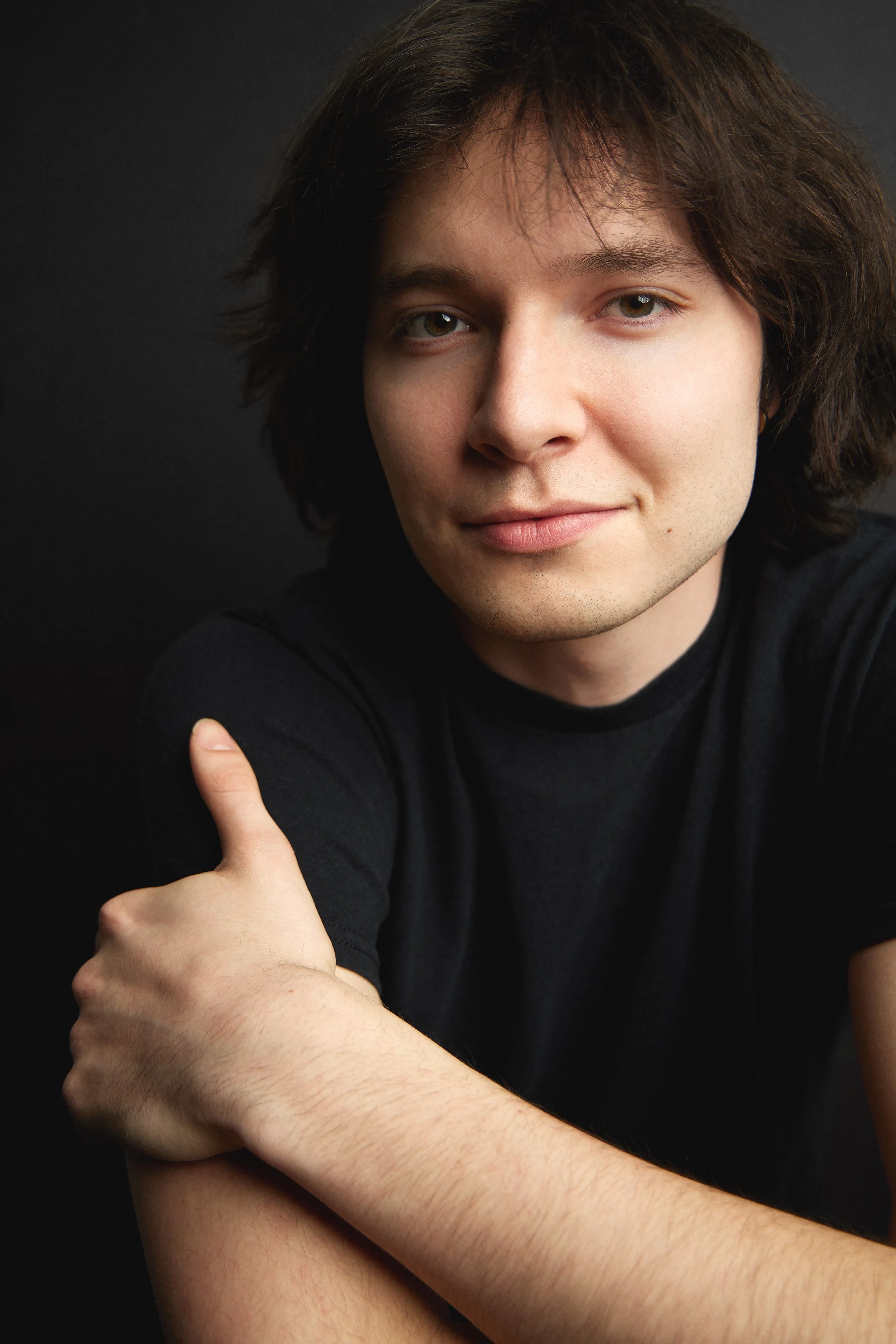 A smiling young man with shaggy brown hair and hazel eyes, wearing a black tee-shirt poses in front of a black background
