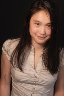 A young Asian woman with brown eyes and black hair wearing a khaki blouse is standing in front of a black background and smiling.