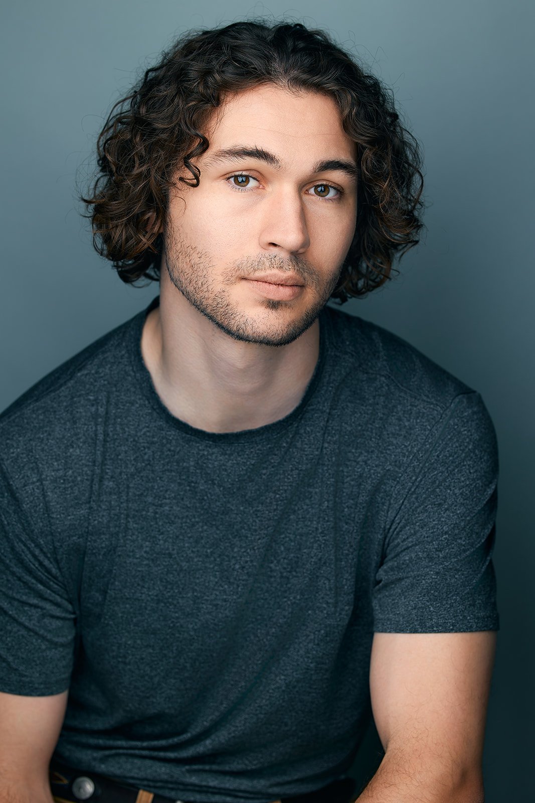 A young man with brown eyes, a close-trimmed beard, and curly, brown, chin-length hair, wearing a gray, heathered tee, sits in front of a neutral background.