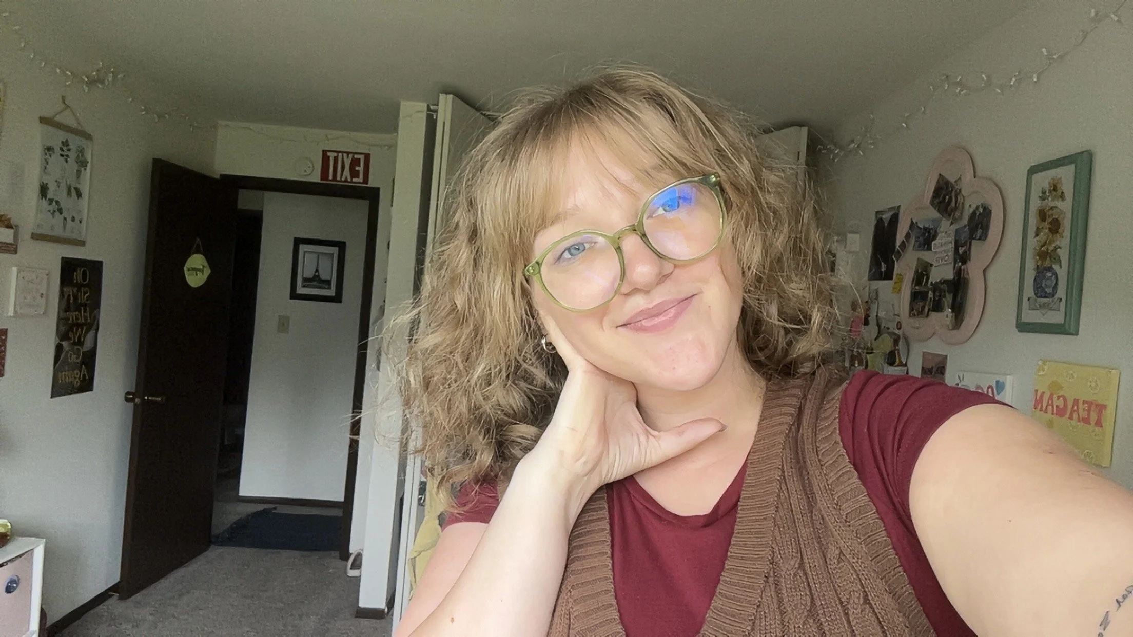 A young woman with a curly blonde bob with bangs, green glasses, and blue eyes, wearing a brown, knitted vest over a red tee shirt is leaning her face against her hand in front of a personal art wall.