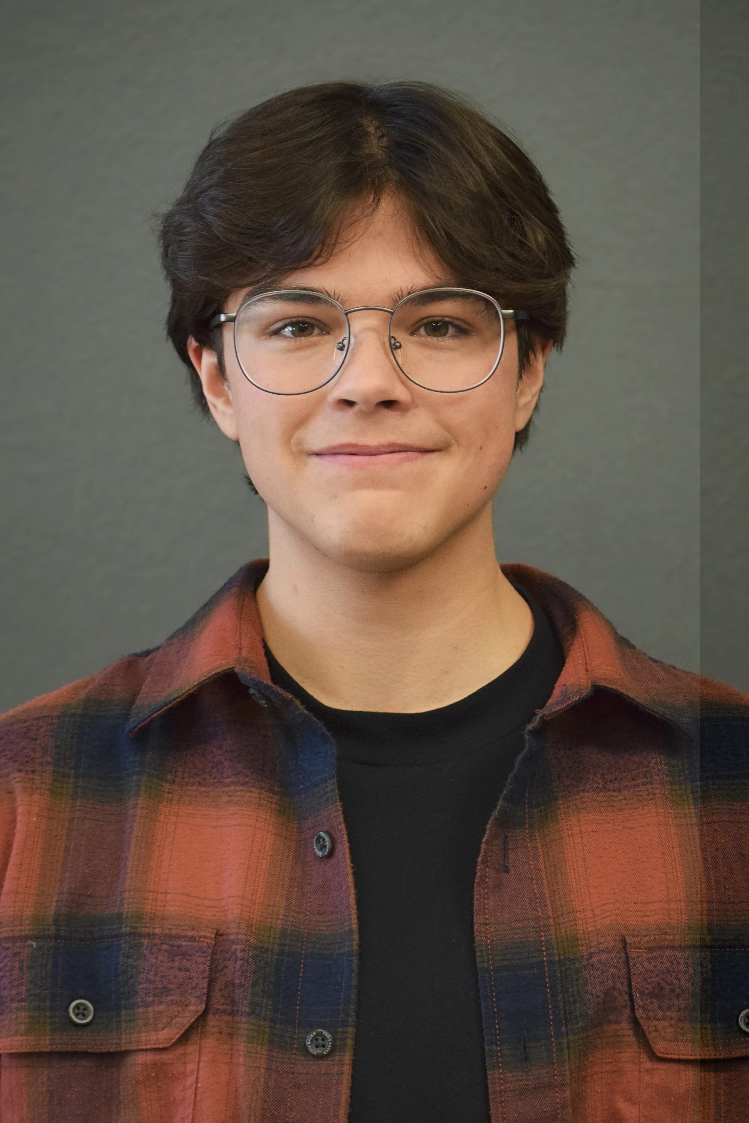 A young man with brown eyes and brown hair with a middle part is wearing a plaid button down over a black tee shirt and glasses.