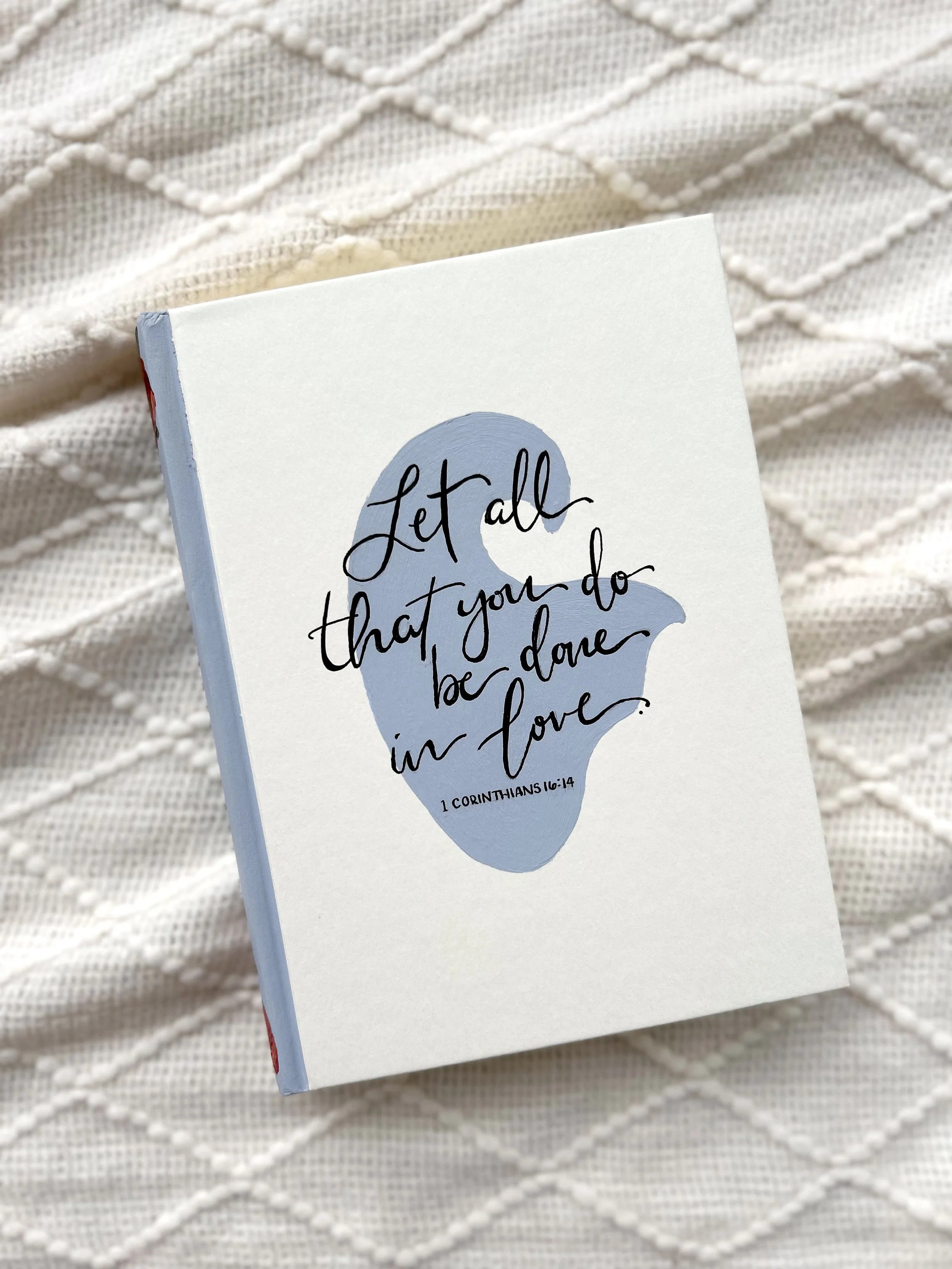 A white book with handwritten text in black ink saying "Let all that you do be done in love" and a small verse reference "1 Corinthians 16:14", placed on a textured white fabric background.