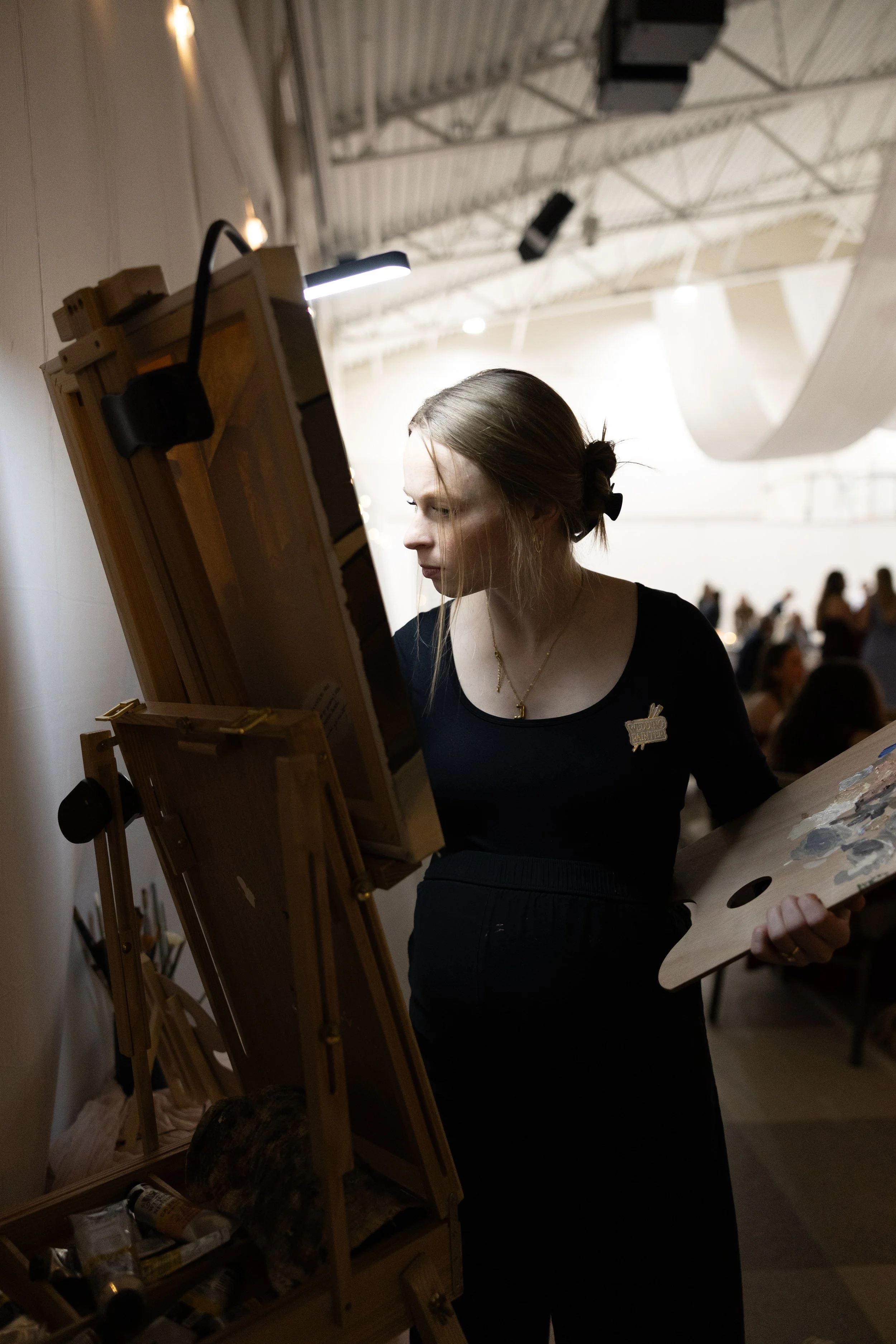 A young woman artist in black clothing, with her hair tied back, standing and painting on a canvas set up on an easel in an indoor art studio or gallery, with other people in the background.