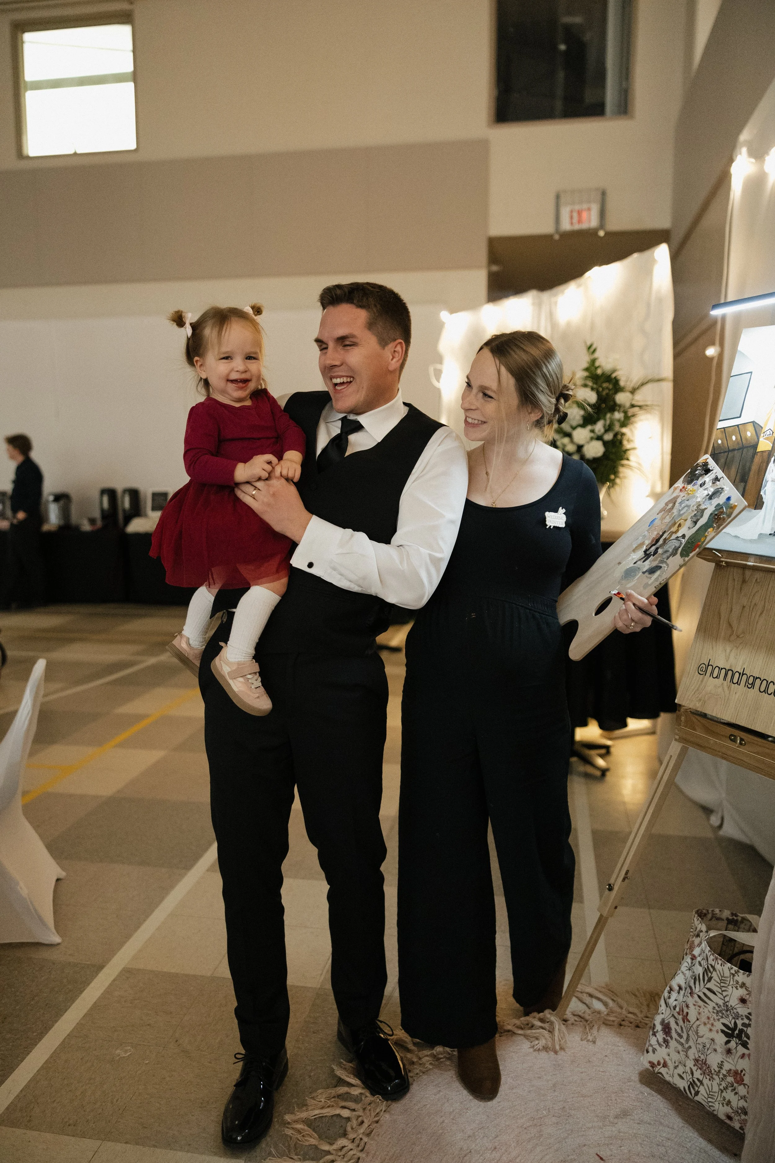 A man holding a young girl in a red dress, with a woman standing beside them, in an indoor event or celebration setting.
