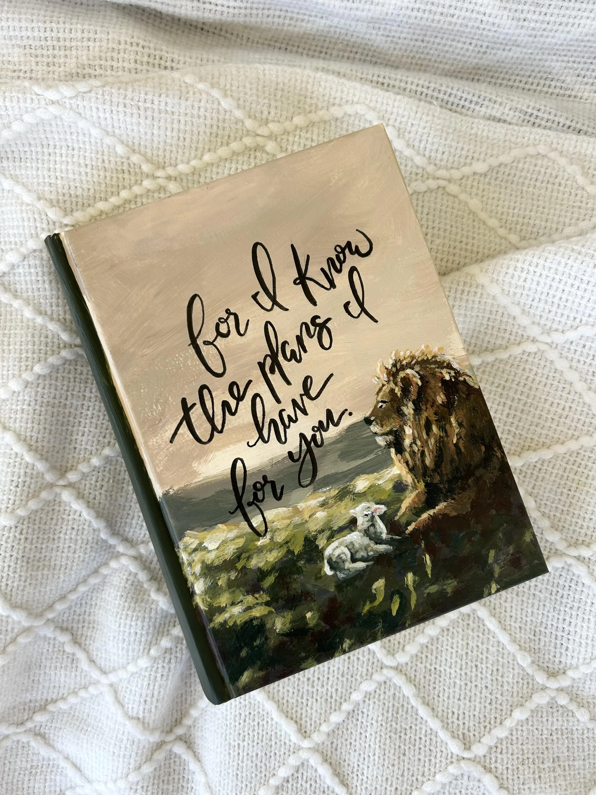 A closed book with a painted cover featuring a lion, sheep, and greenery, placed on a textured white knit blanket.