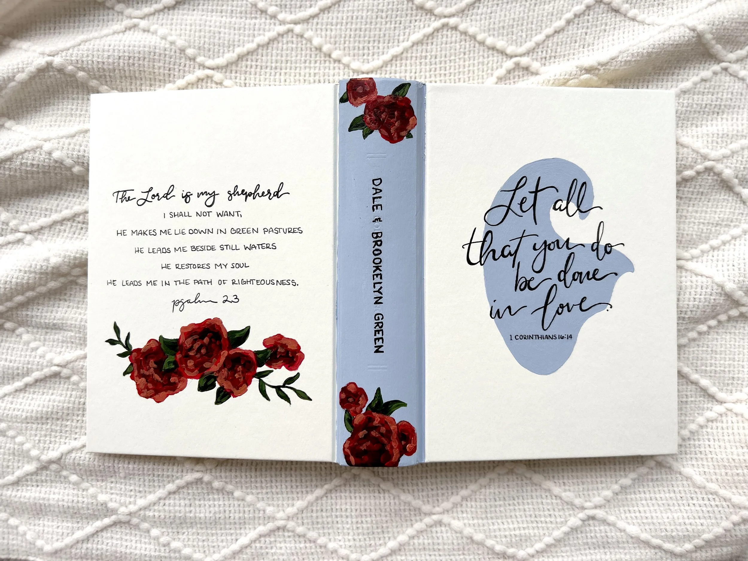 Open greeting card with red roses and greenery on the left and a blue abstract shape with handwritten-style text on the right, placed on a textured white fabric background.