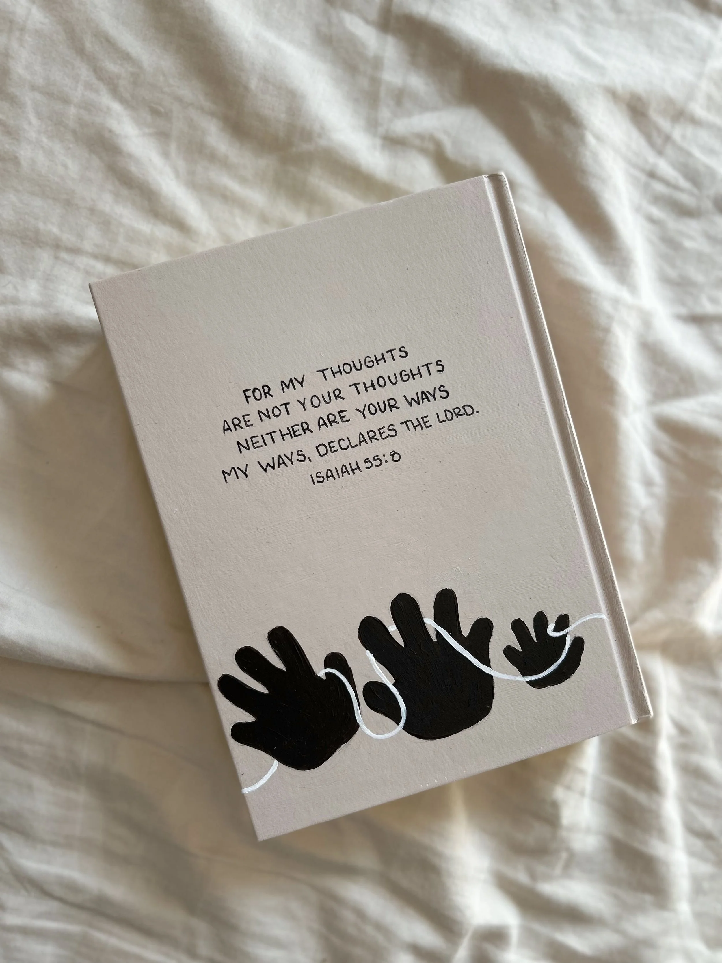Close-up of a white notebook with black and white hand illustrations on the cover, placed on a wrinkled white fabric surface.