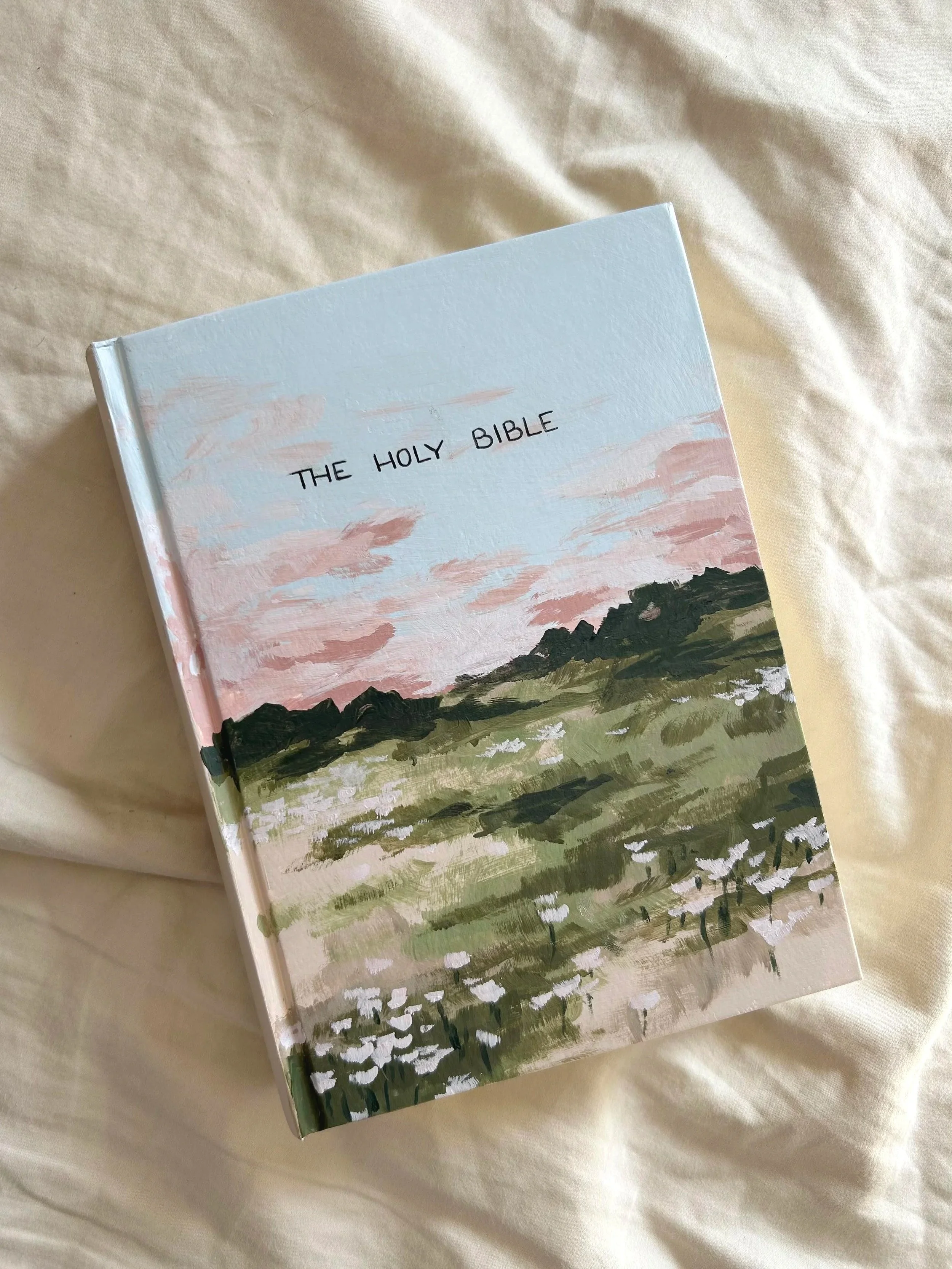 Close-up of a painted book cover titled "The Holy Bible" with pastel-colored clouds and a landscape of trees and hills, placed on a beige fabric surface.